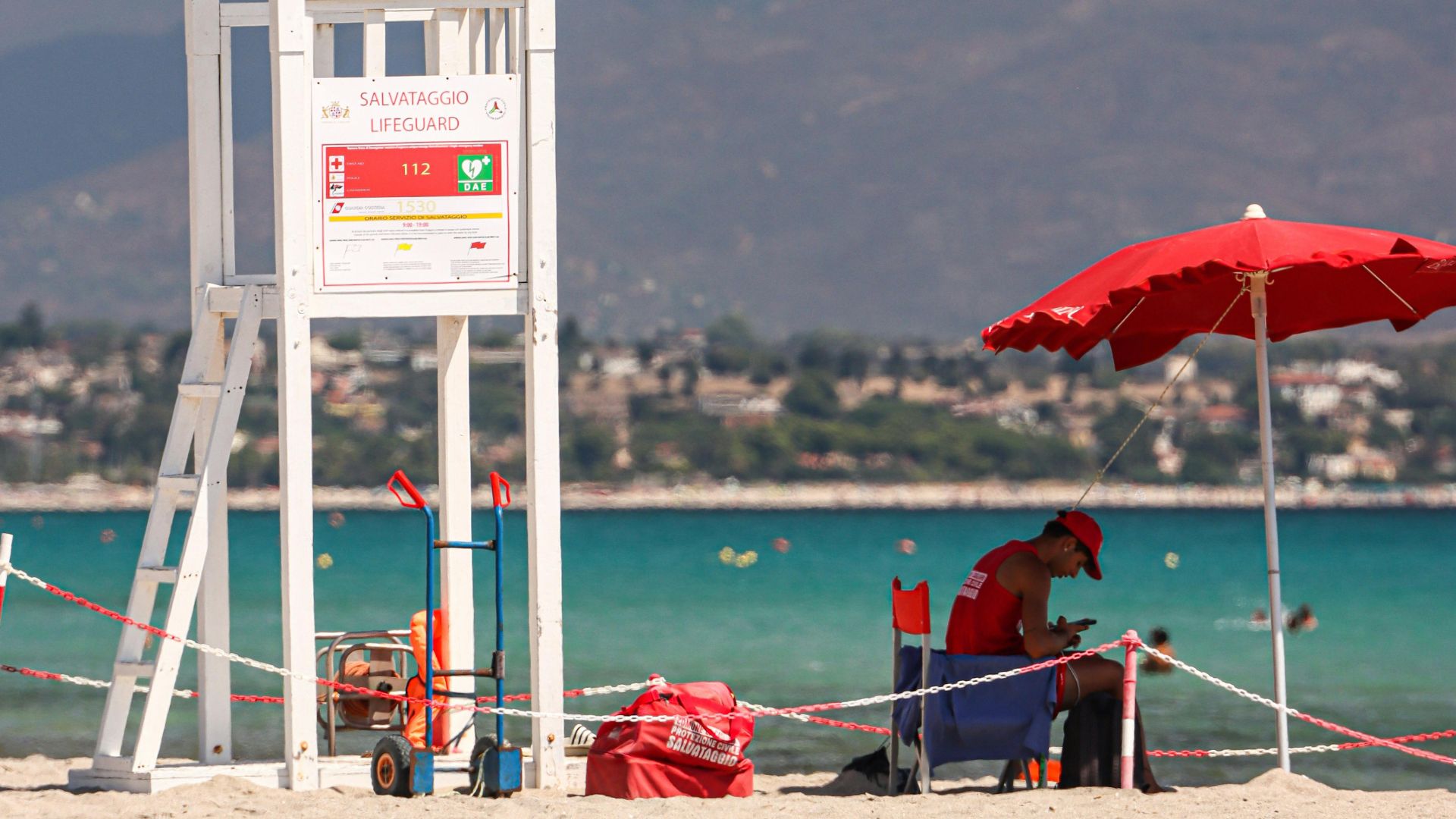 A lifeguard sits under a red umbrella on the sandy Poetto Beach in Cagliari, with a lifeguard station and the coastline visible in the background.