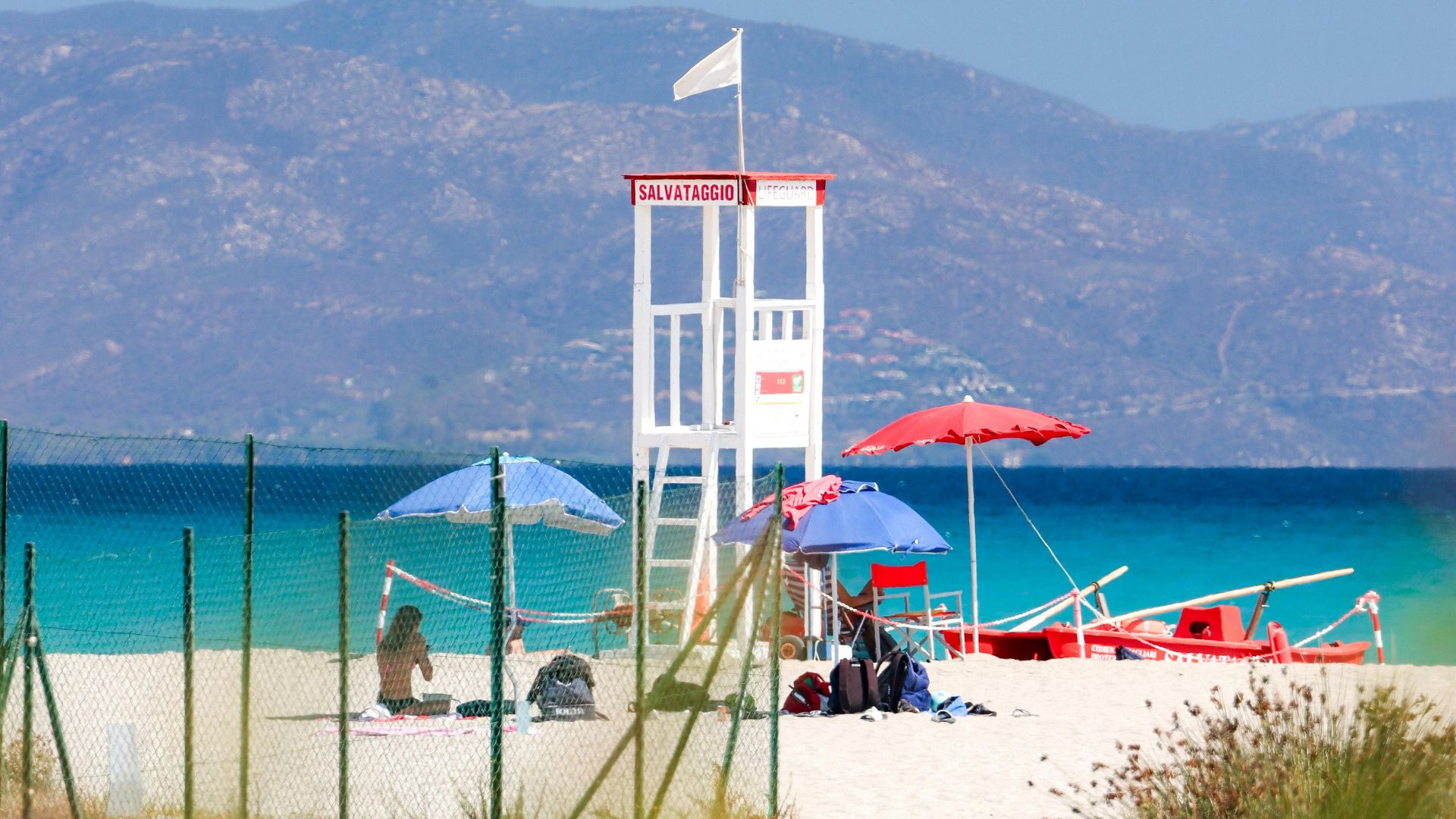 A lifeguard tower and beach umbrellas dot the sandy expanse of Poetto Beach in Cagliari, Sardinia, with the calm, blue sea and distant mountains providing a scenic backdrop.