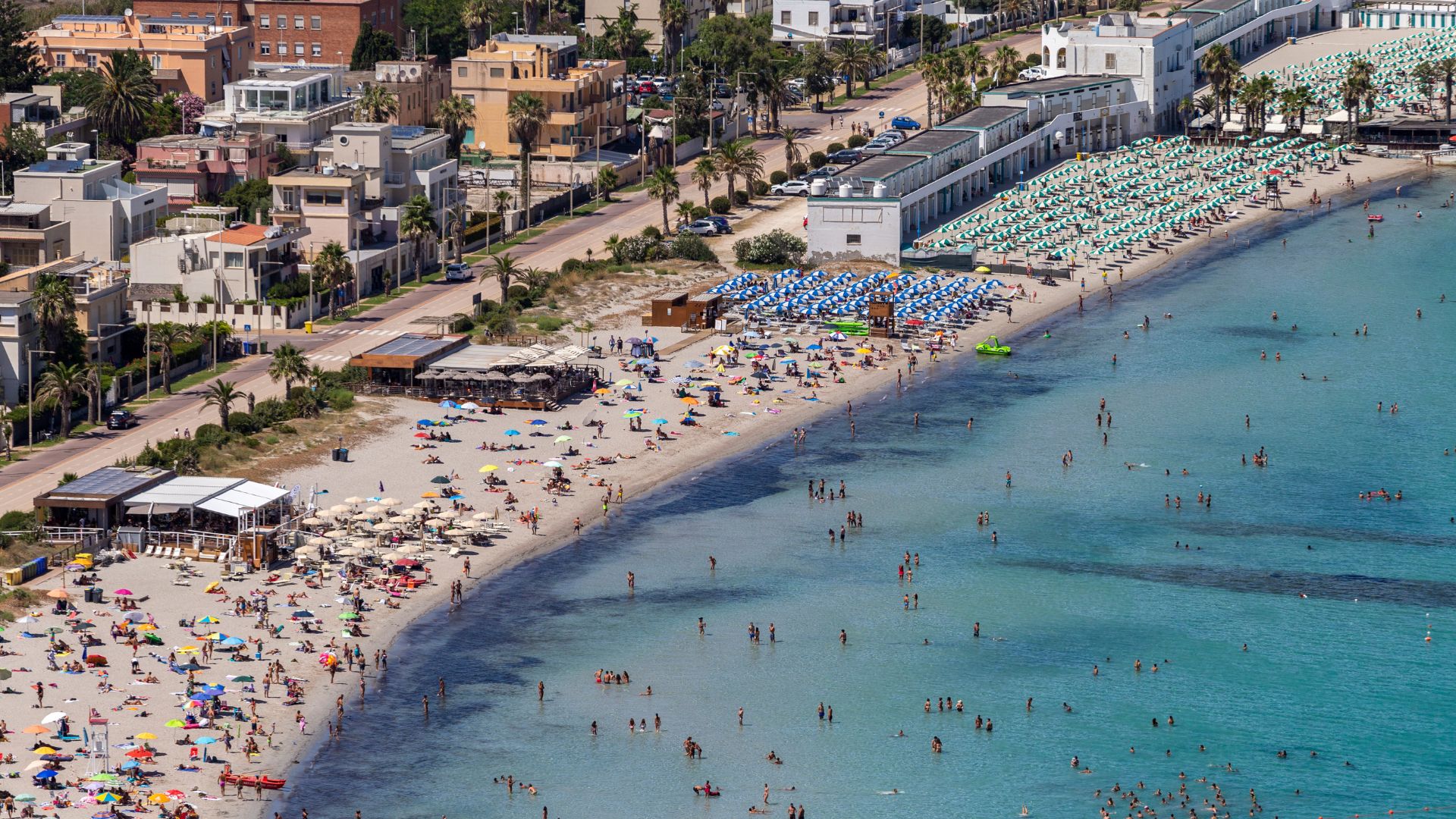 Aerial view of Poetto Beach in Cagliari, Sardinia, showing a crowded beach with colorful umbrellas, clear blue water with swimmers, and the cityscape in the background.