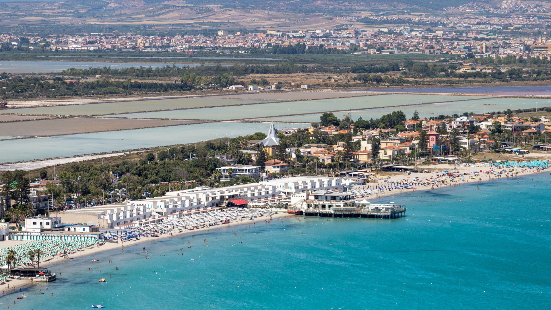 A high-angle view captures the expansive Poetto Beach in Cagliari, Sardinia, featuring a long stretch of white sand bordered by the turquoise waters of the Mediterranean Sea. The beach is dotted with colorful umbrellas and sunbathers, while a line of buildings and a marina structure extend along the shoreline. In the background, the city of Cagliari is visible, nestled against a backdrop of hills and a lagoon.