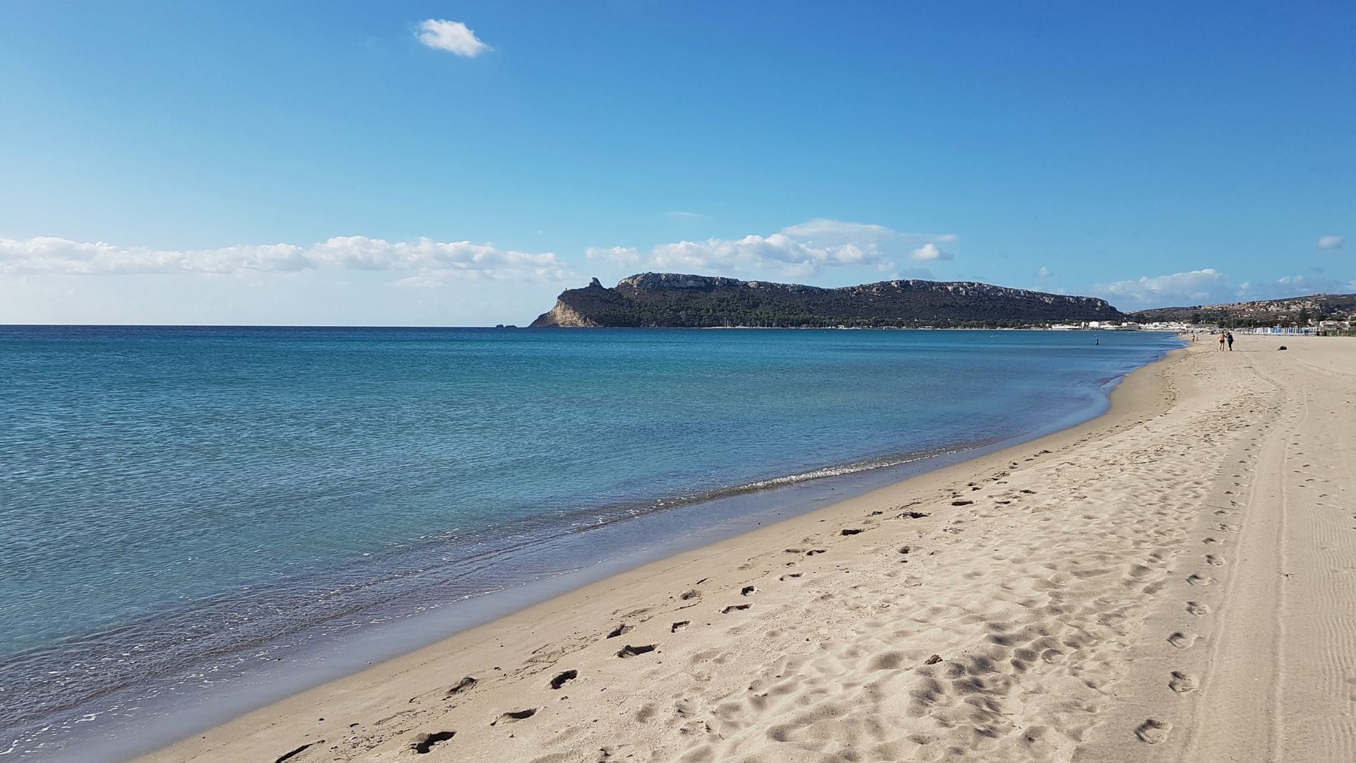 A scenic view of Poetto Beach in Cagliari, Sardinia, featuring a long stretch of sandy shore meeting the clear blue waters of the Mediterranean Sea, with the silhouette of Sella del Diavolo visible in the background under a clear sky.