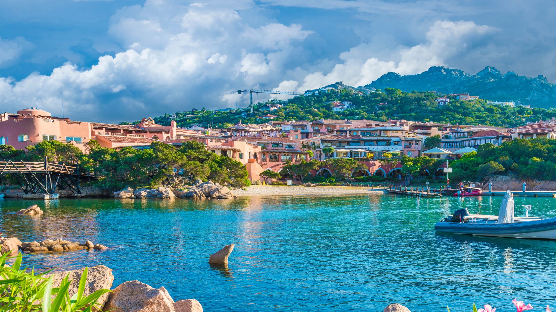 A coastal view of Porto Cervo, Sardinia, featuring colorful buildings, lush greenery, and a clear blue sea under a partly cloudy sky.