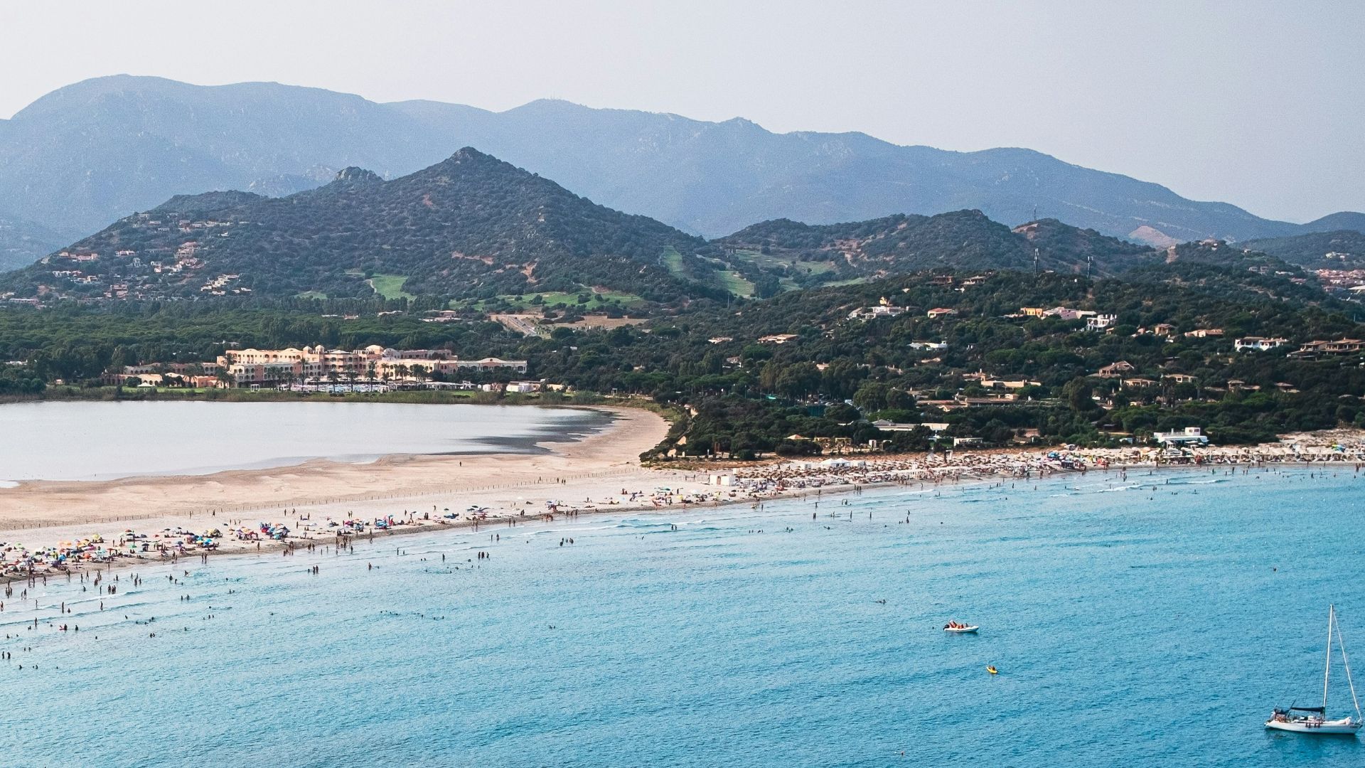 A panoramic view of Spiaggia di Porto Giunco in Sardinia, featuring a crowded beach, blue sea, lagoon, and mountains.
