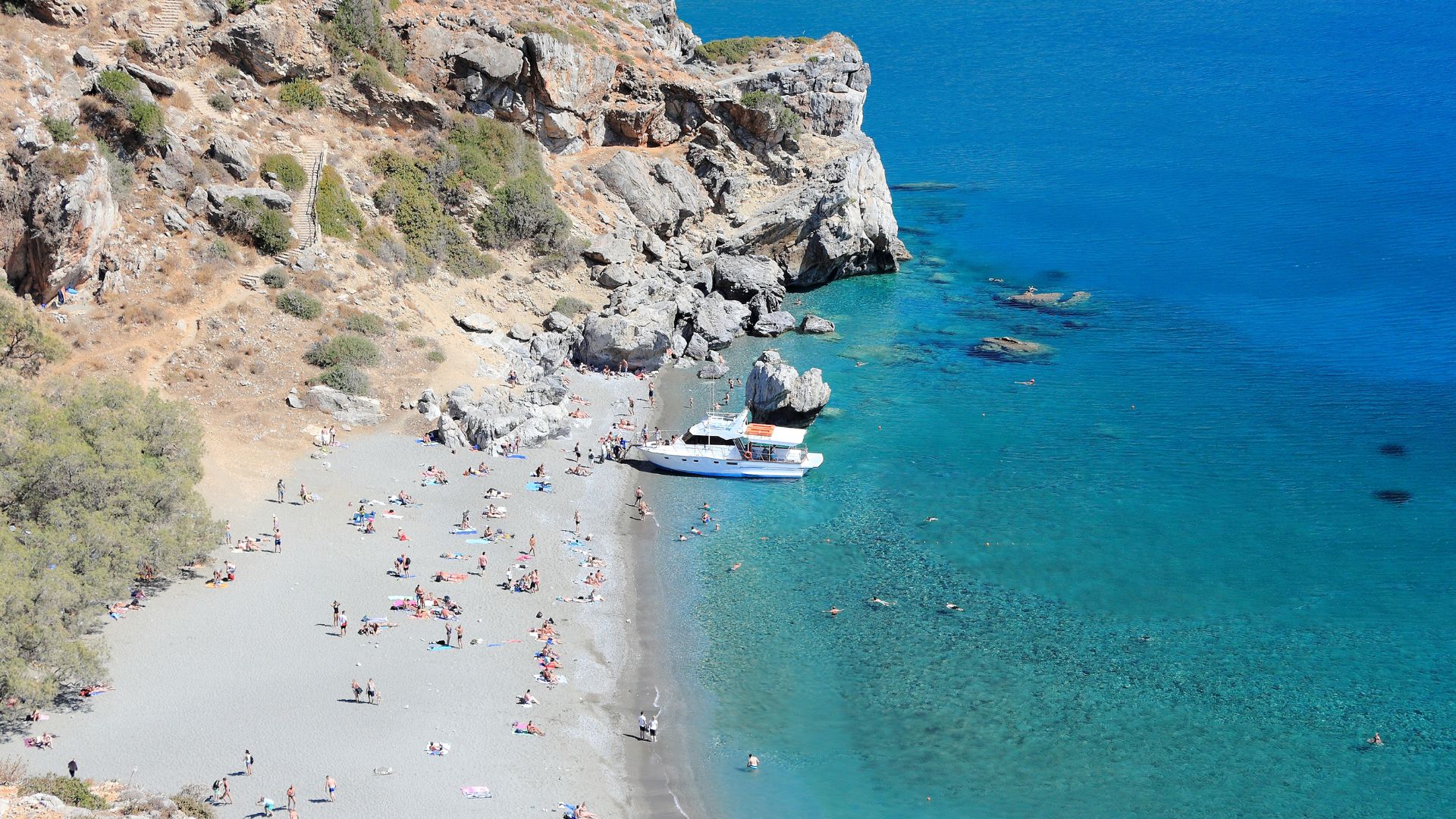 Aerial view of Preveli Beach in Crete, Greece, featuring a sandy beach, turquoise sea, a river flowing into the sea, palm trees, and rocky cliffs.