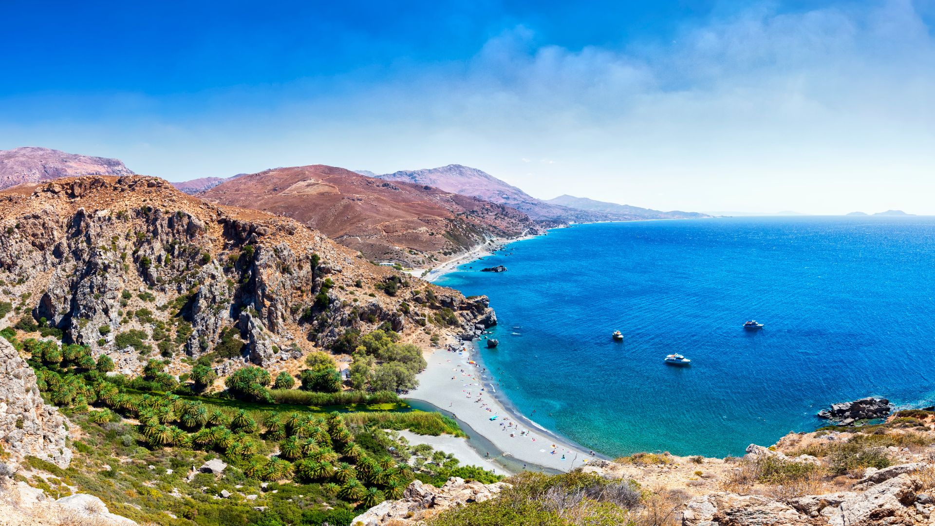Aerial view of Preveli Beach in Crete, Greece, featuring a sandy beach, turquoise sea, a river flowing into the sea, palm trees, and rocky cliffs.