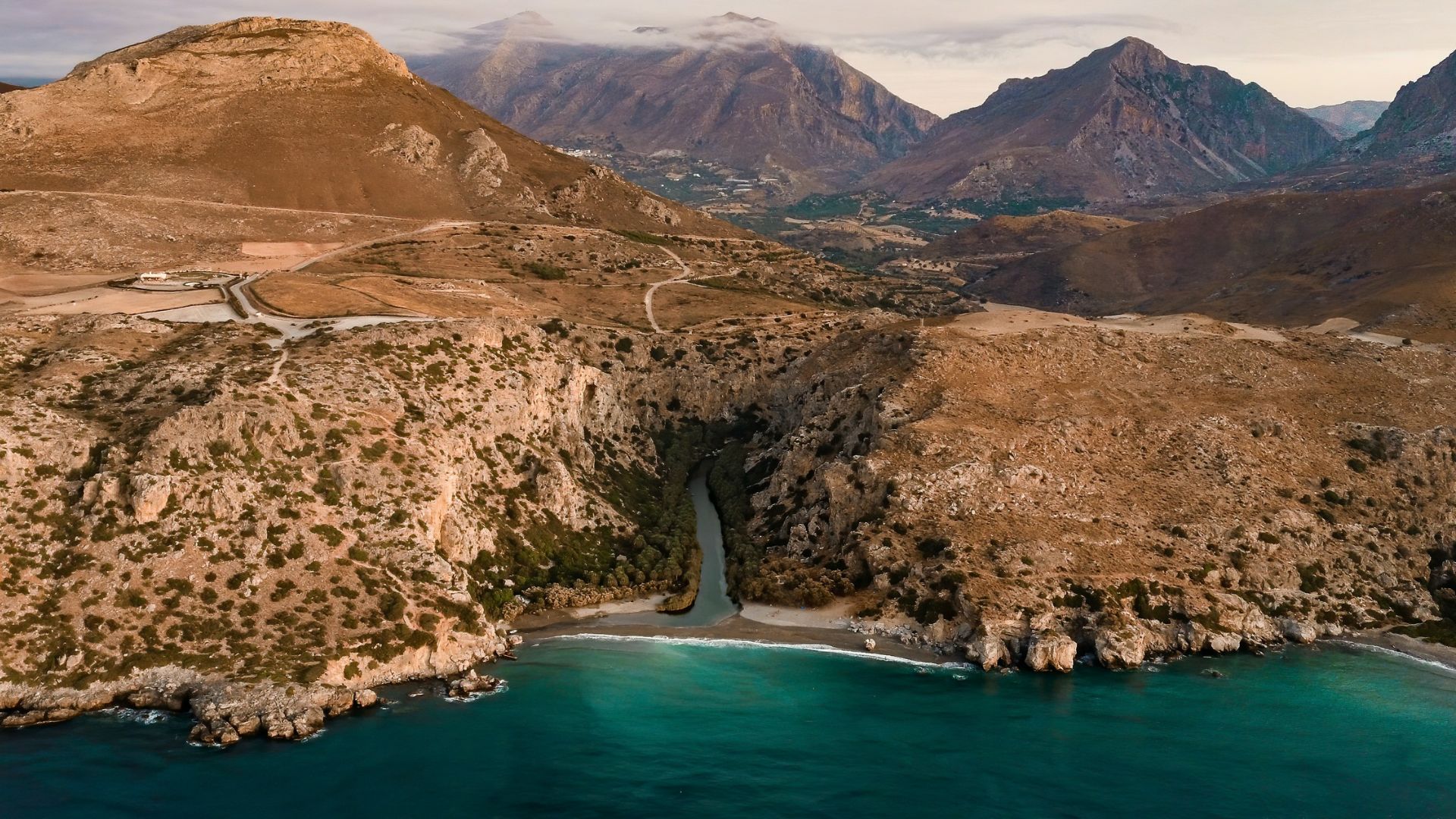 Aerial view of Preveli Beach, where Megas Potamos River flows into the sea, flanked by palm trees and rocky cliffs, with mountains in the background.