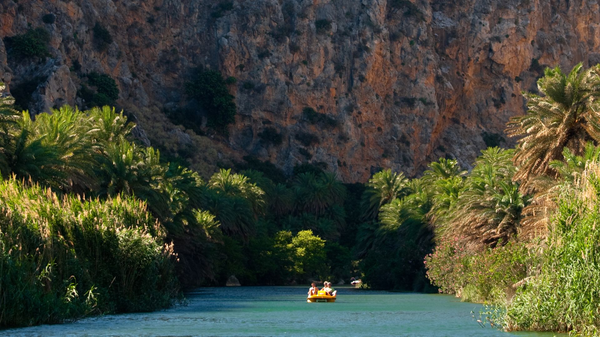 A river flows through a palm forest towards Preveli Beach, with a rocky cliff face in the background and a small boat on the water.