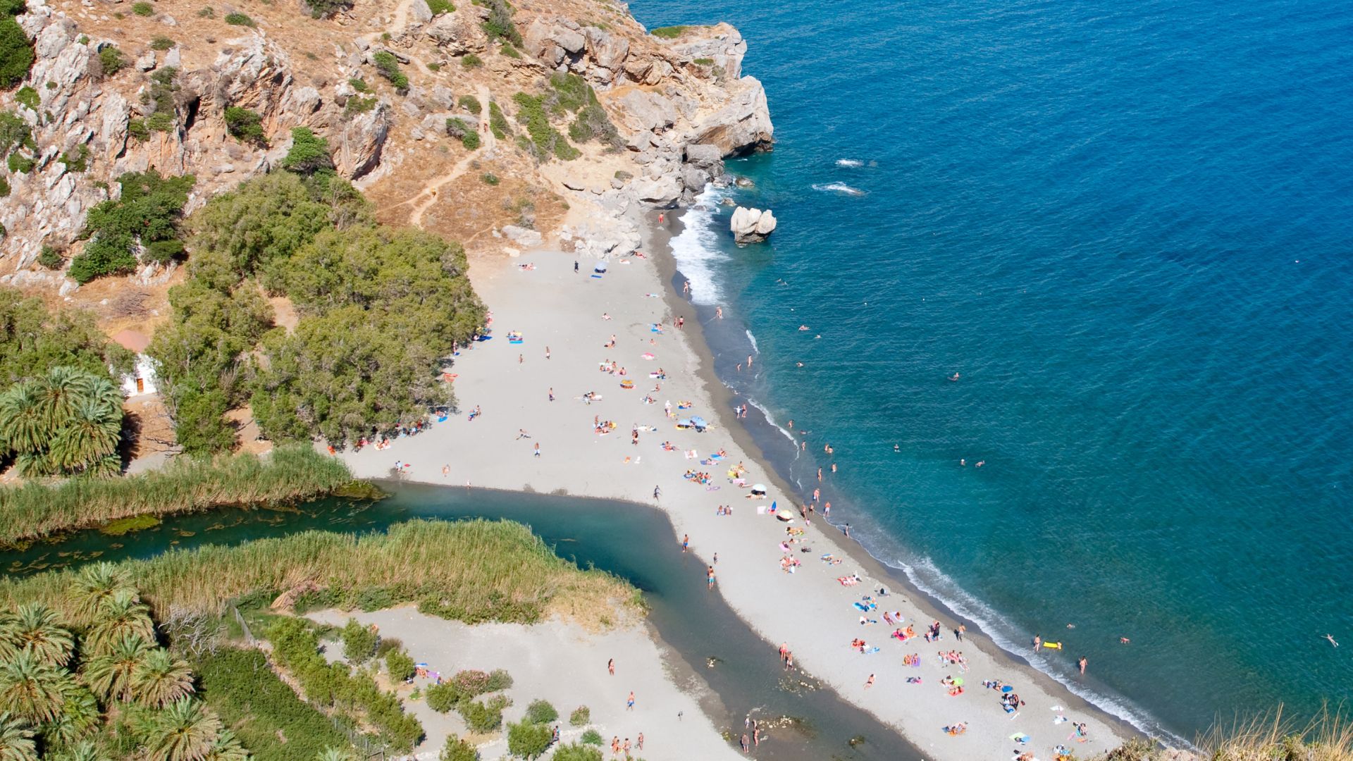Aerial view of Preveli Beach in Crete, Greece, featuring a sandy beach, turquoise sea, a river flowing into the sea, palm trees, and rocky cliffs.