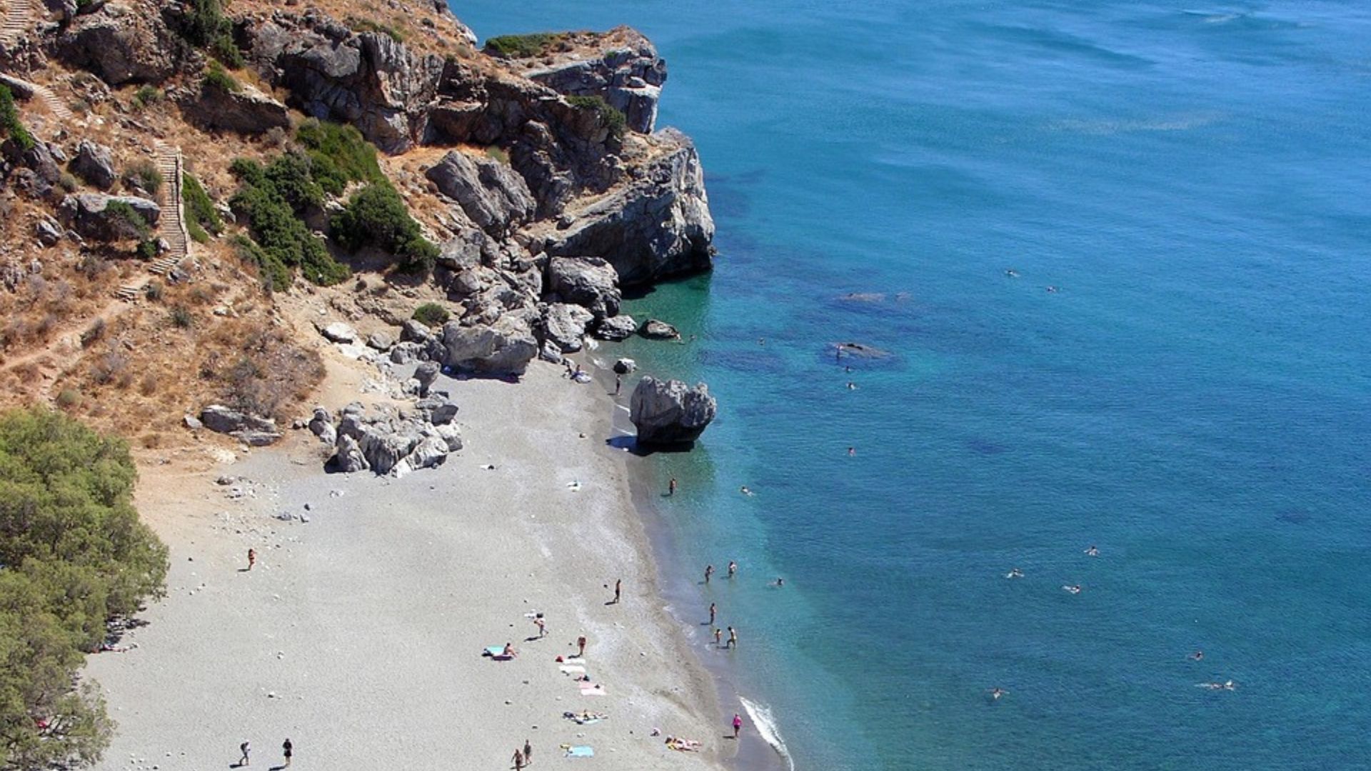 Aerial view of Preveli Beach in Crete, featuring a sandy beach, a river flowing into the sea, rocky cliffs, and the surrounding blue waters of the Mediterranean.