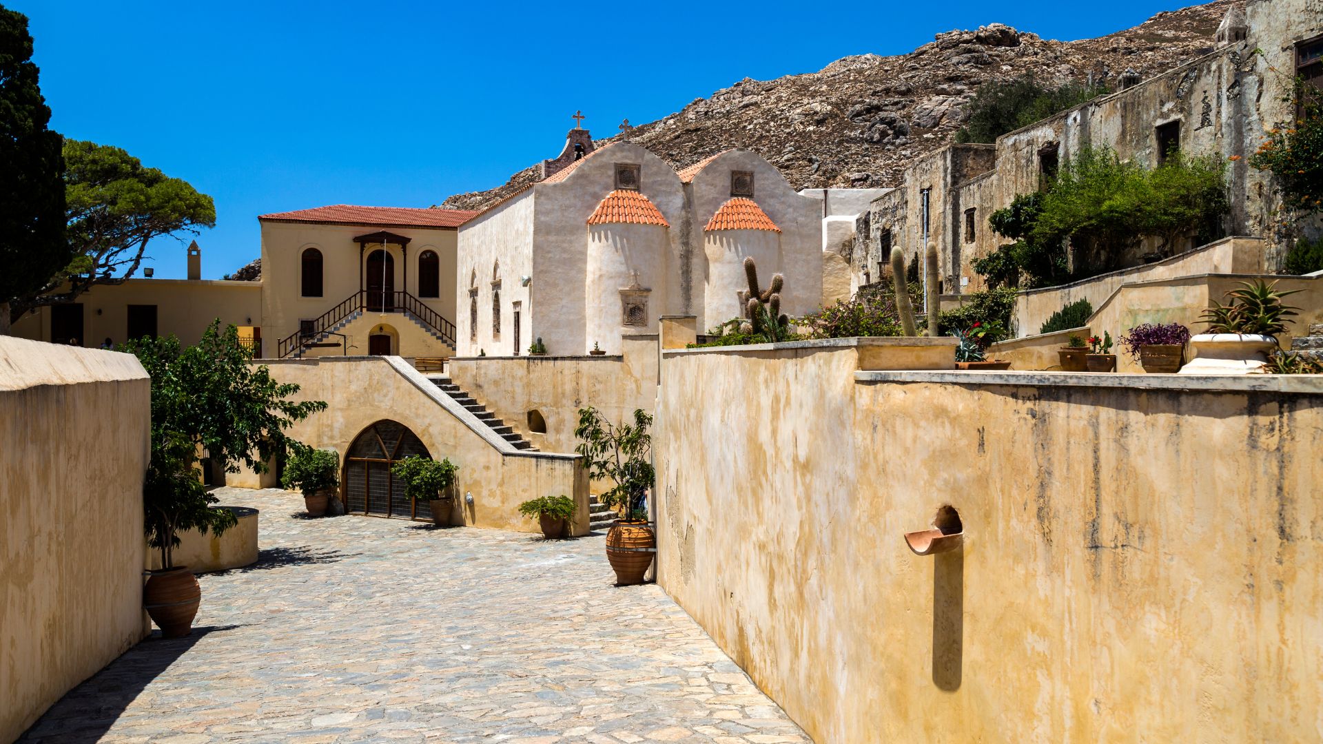 Exterior view of the historic Preveli Monastery complex in Crete, featuring stone buildings, a courtyard, and surrounding landscape.