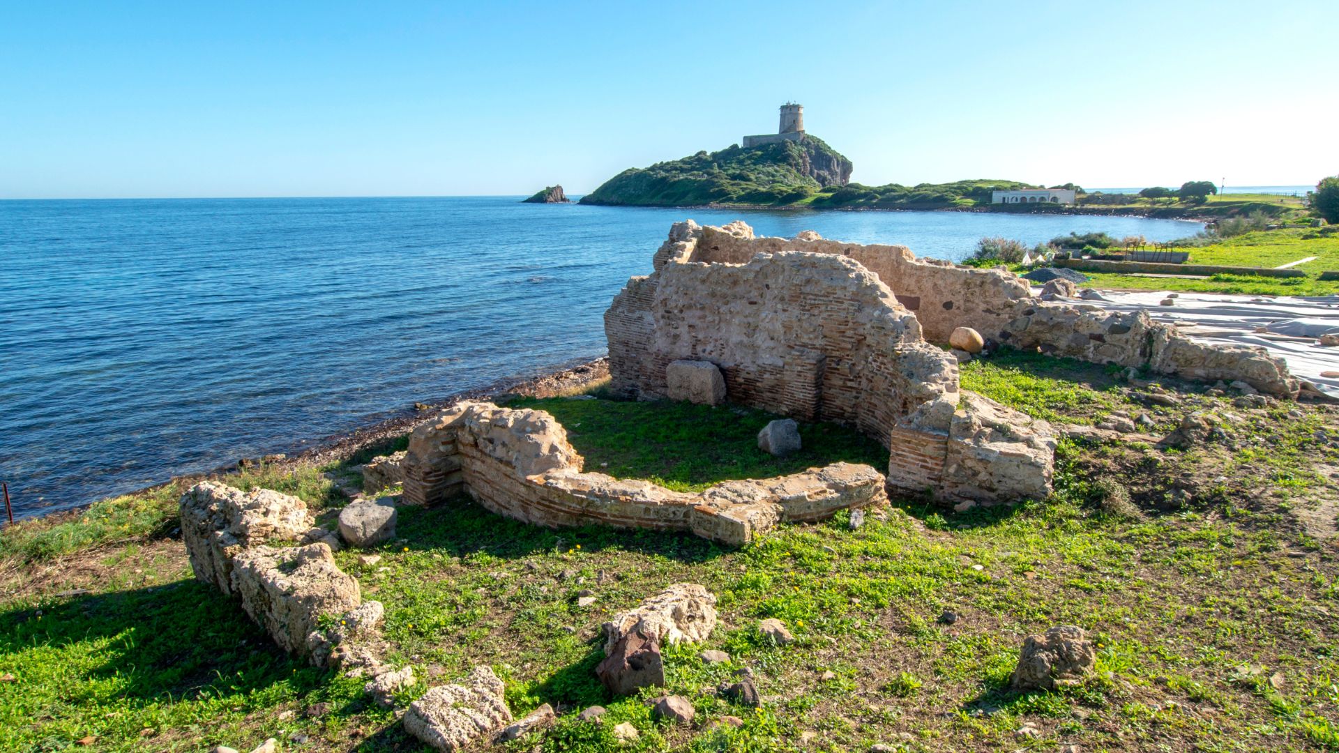 Ruins of Nora, an ancient Roman city on the coast of Sardinia, with a tower visible in the background. The ruins consist of stone walls and foundations, surrounded by grass and the sea. 