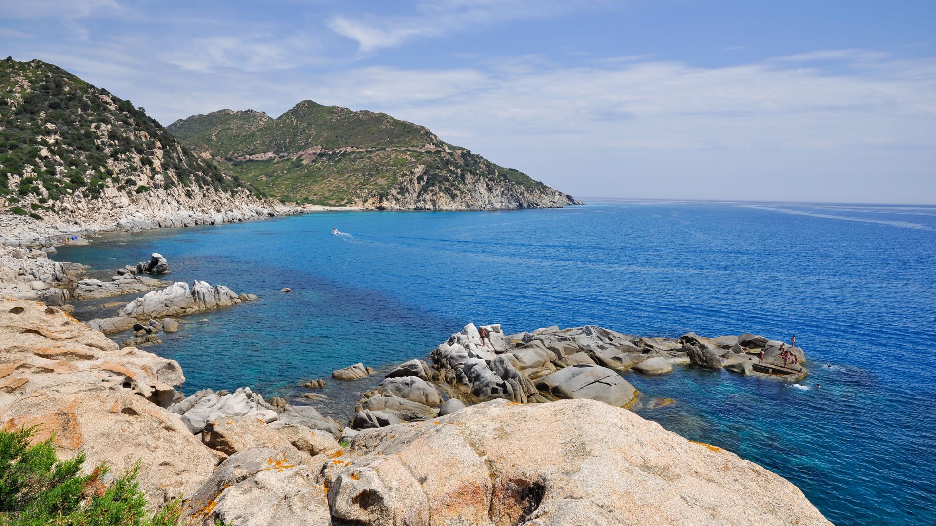 A scenic view of Punta Molentis Beach in Sardinia, featuring clear turquoise water, smooth granite rocks, and a mountainous coastline in the background.