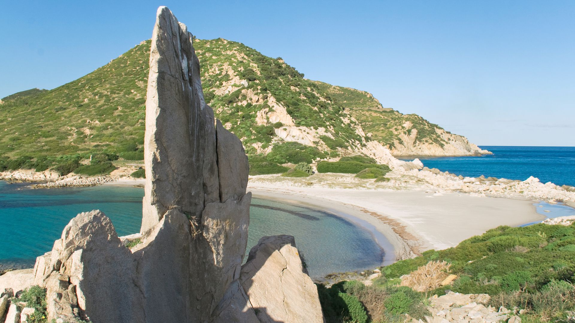 A striking rock formation juts out against the backdrop of Punta Molentis Beach in Sardinia, with its sandy shore and clear blue waters.