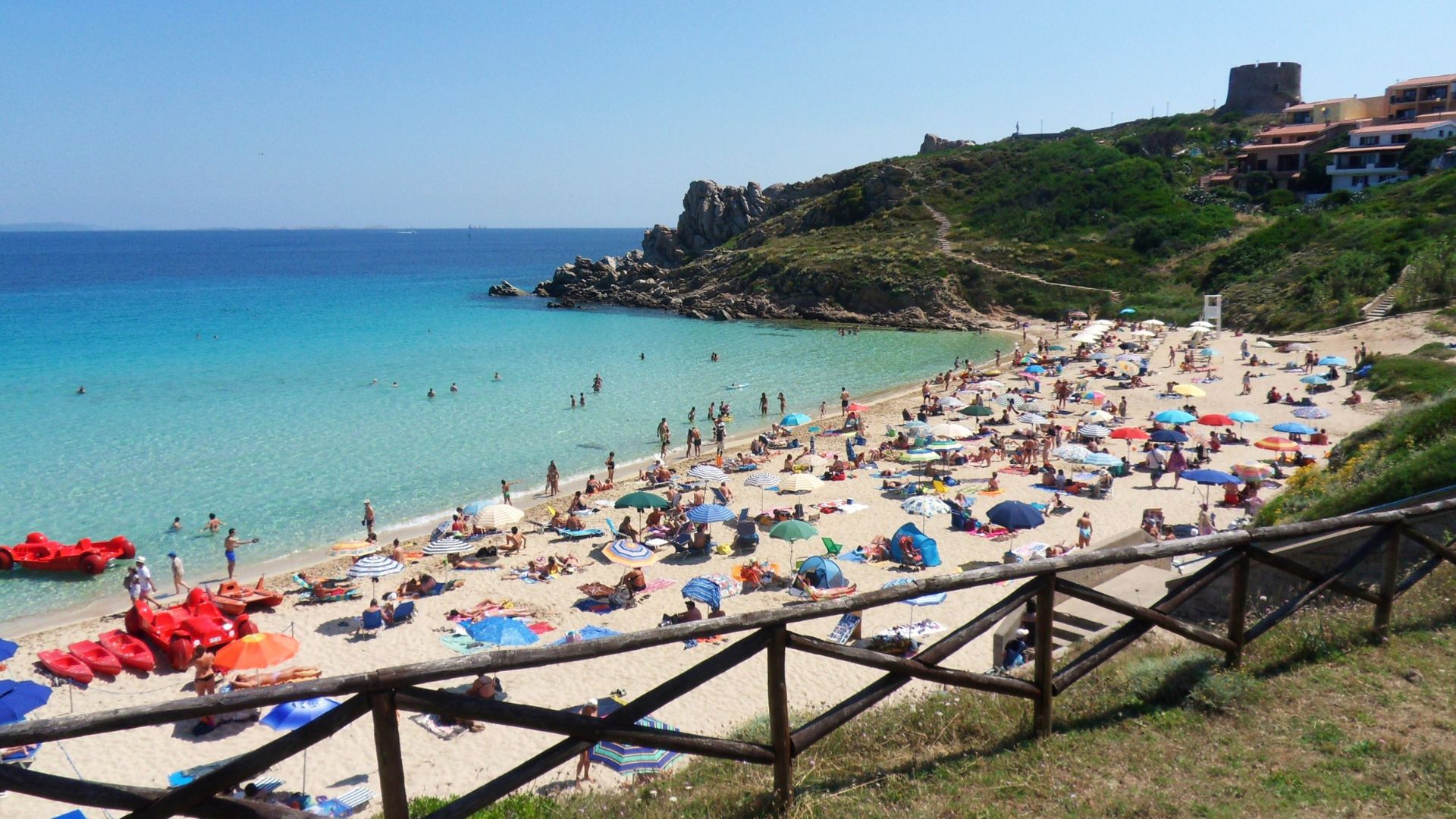 A scenic view of Rena Bianca beach in Sardinia, Italy, featuring white sand, clear turquoise water, beachgoers, umbrellas, and a rocky coastline with a tower in the background.