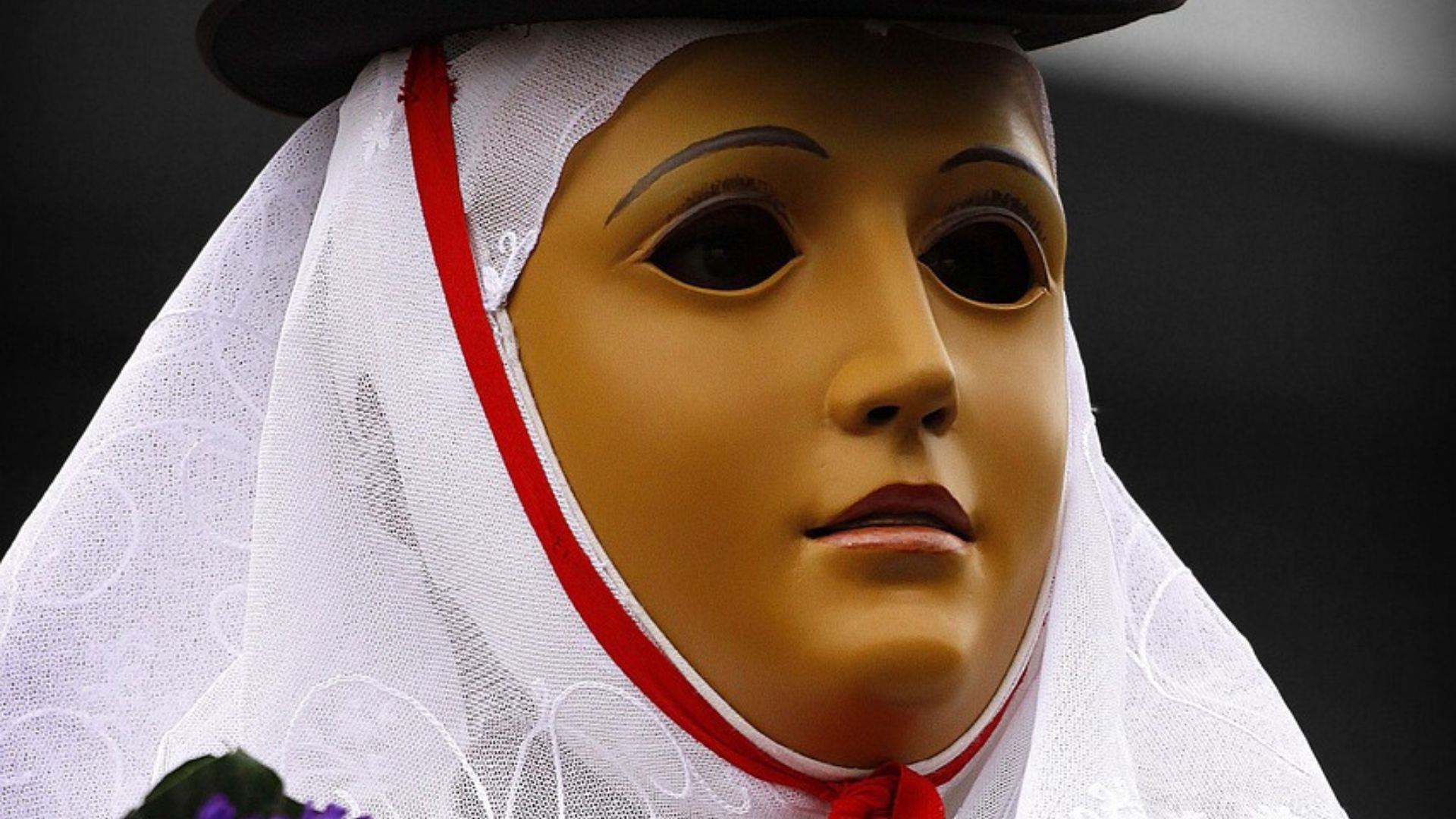 A close-up of a mask, possibly used in the Sartiglia of Oristano, featuring a pale yellow face, black eyes, and red-outlined white veil. The mask is framed by a black hat and set against a dark background. 