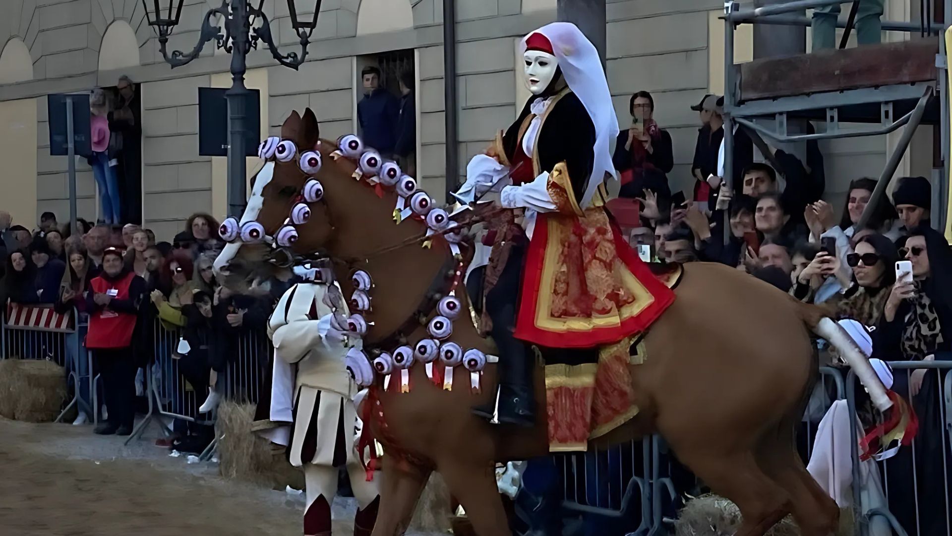 Su Componidori, dressed in traditional attire with a white mask and ornate costume, rides a decorated horse during the Sa Sartiglia festival, surrounded by spectators
