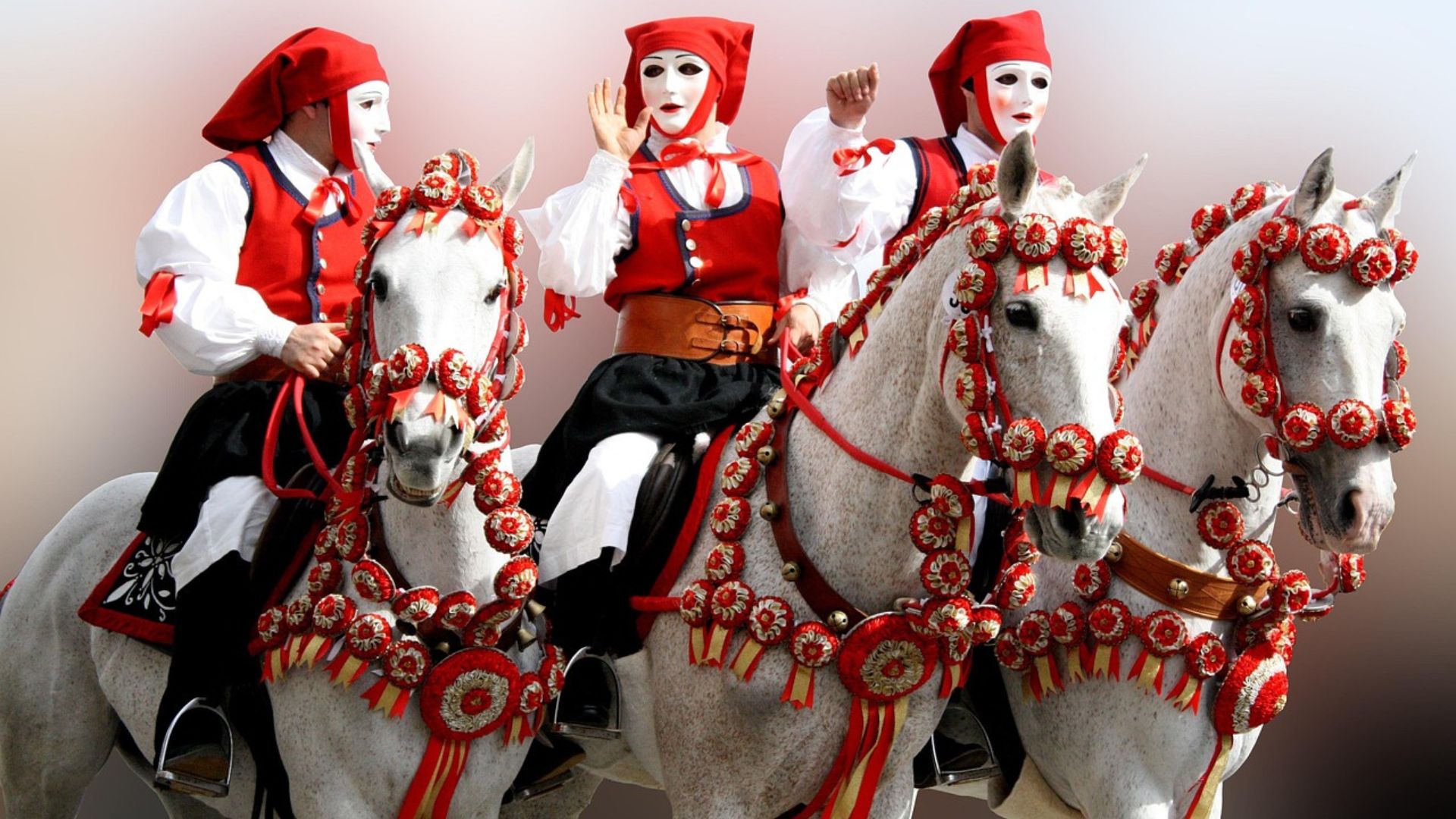 Three riders in traditional costumes and masks on decorated white horses during Sartiglia. The horses are adorned with red and white decorations, while the riders wear red and white outfits with masks, participating in the traditional Sardinian equestrian event.