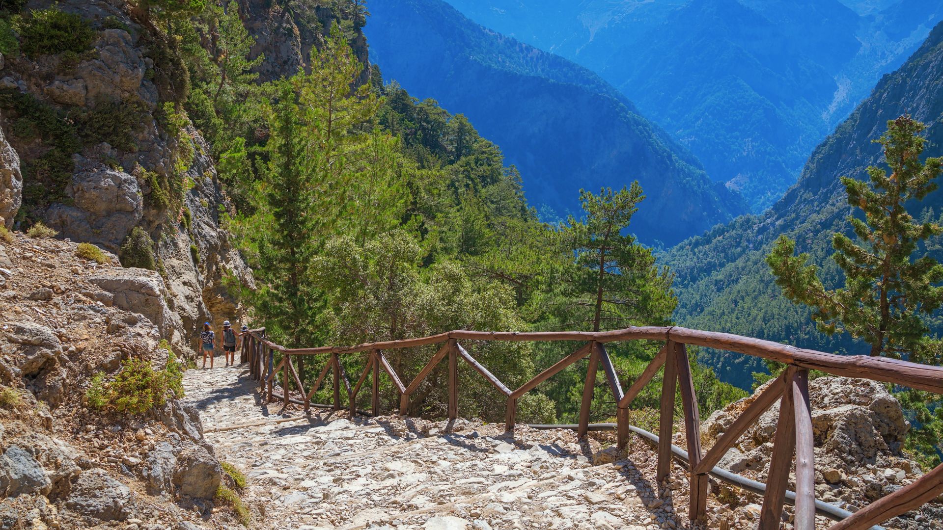 Samaria Gorge trail in Samaria National Park, Crete, featuring a stone path with a wooden railing, surrounded by steep, rocky cliffs and lush vegetation, with distant blue mountains in the background. 
