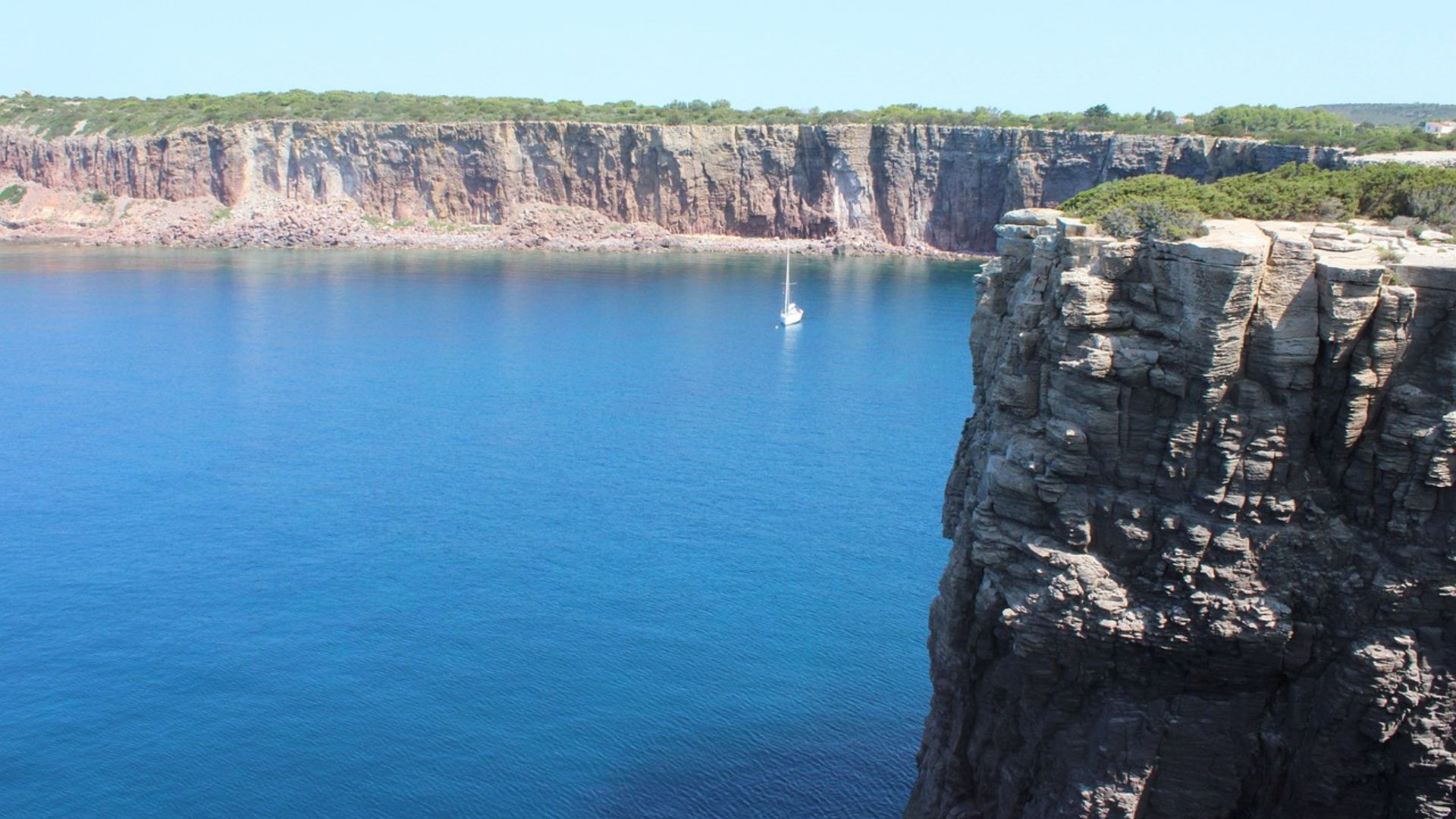 Blue waters, a sailboat, steep rocky cliifs of San Pietro Island (Isola di San Pietro), Carloforte, Sardinia, Italy.