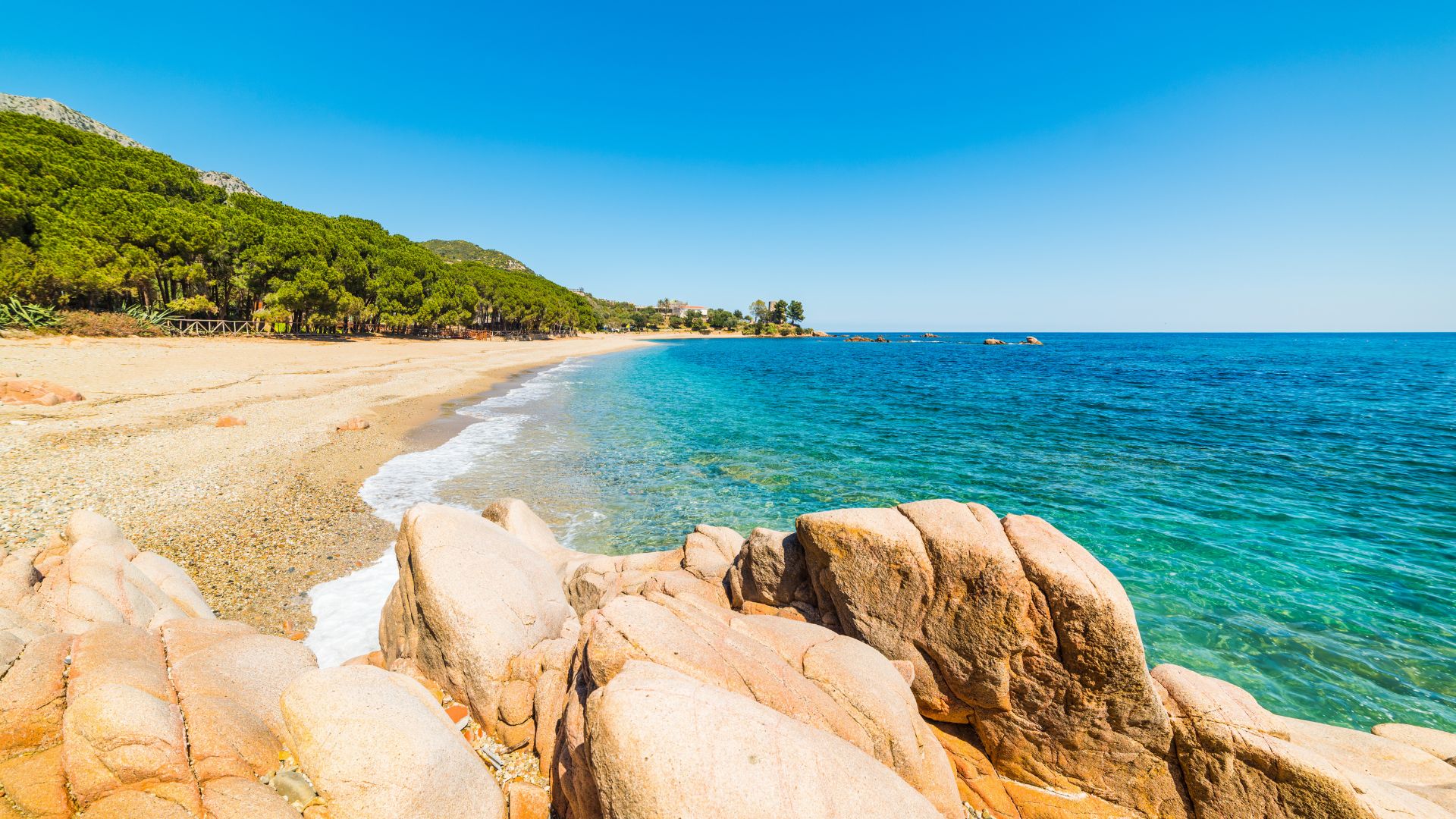 A scenic view of Spiaggia di Santa Maria Navarrese in Sardinia, featuring a sandy beach meeting turquoise waters, with rocky formations in the foreground and a green hillside in the background under a clear blue sky.