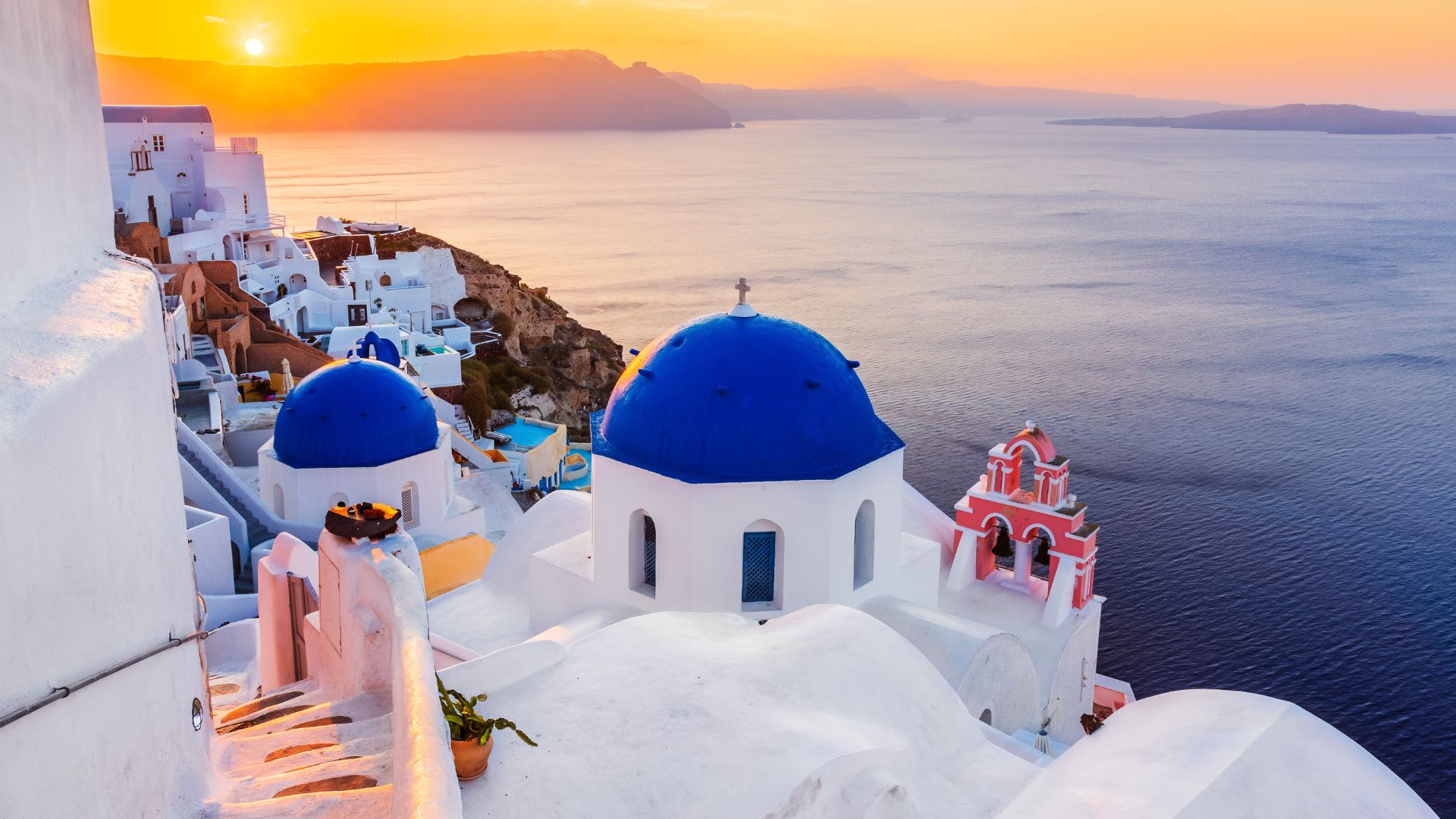A scenic sunset view of Santorini with its iconic white buildings and blue domes overlooking the Aegean Sea.