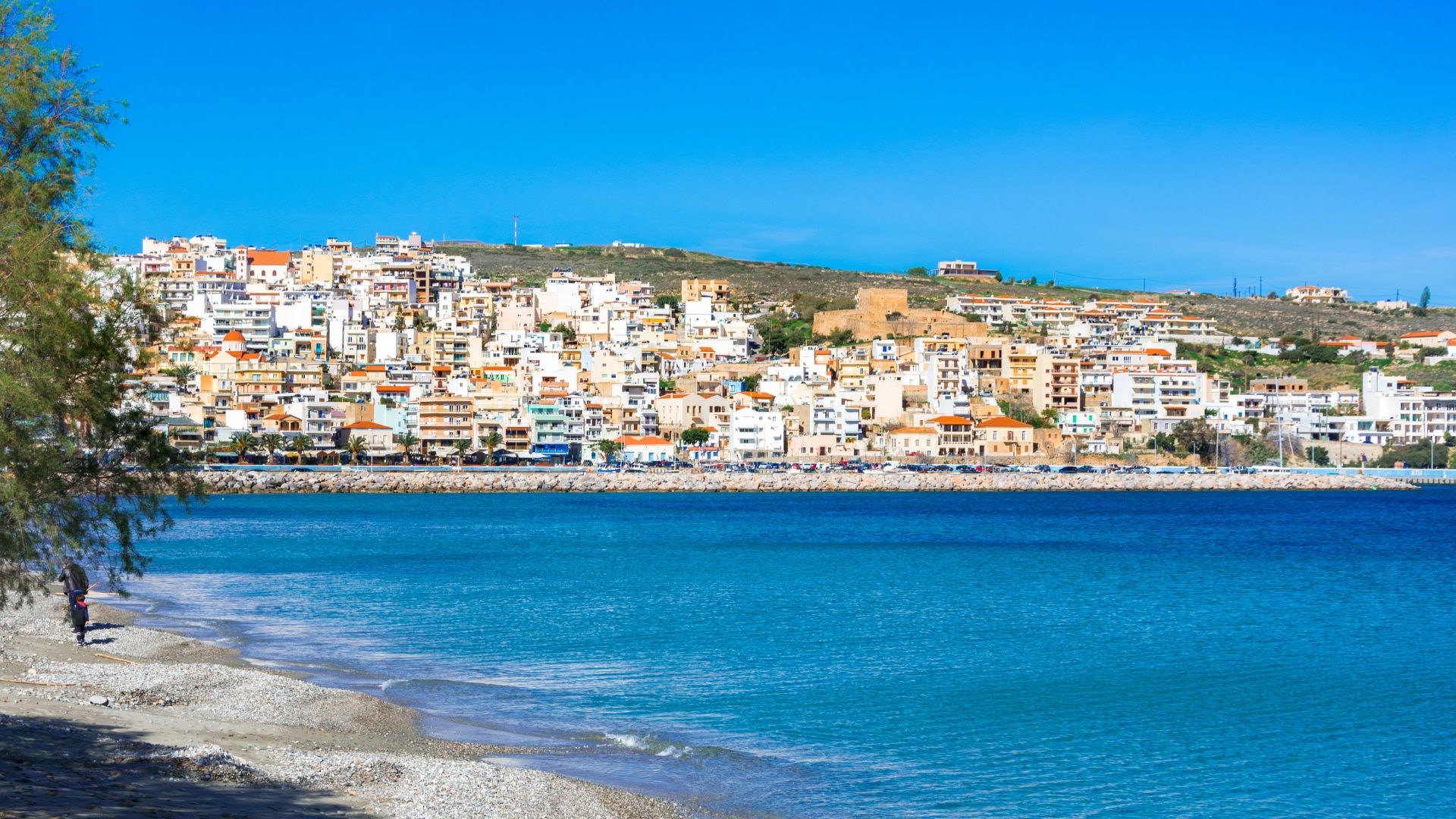 A coastal view of Sitia Beach, Crete, with a town on a hillside and turquoise waters.