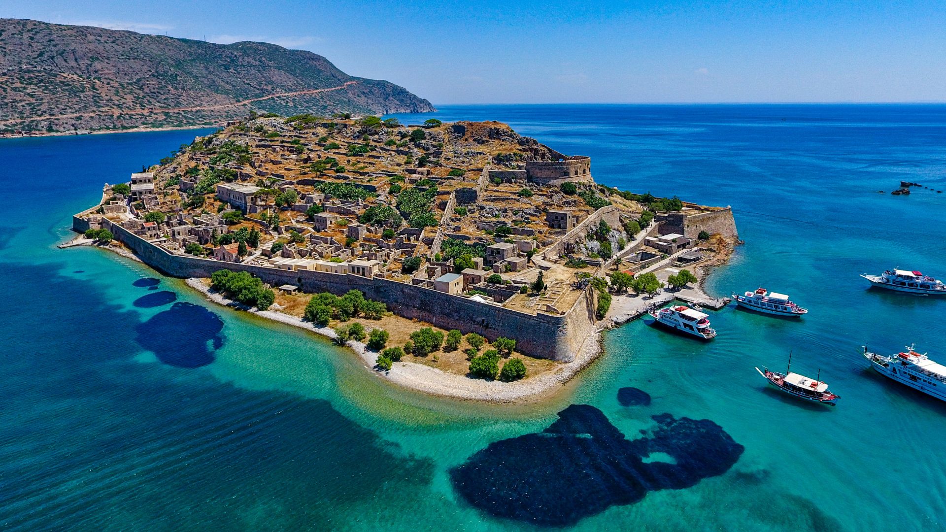 Aerial view of Spinalonga Island, Crete, featuring the Venetian fortress ruins surrounded by turquoise waters and boats.