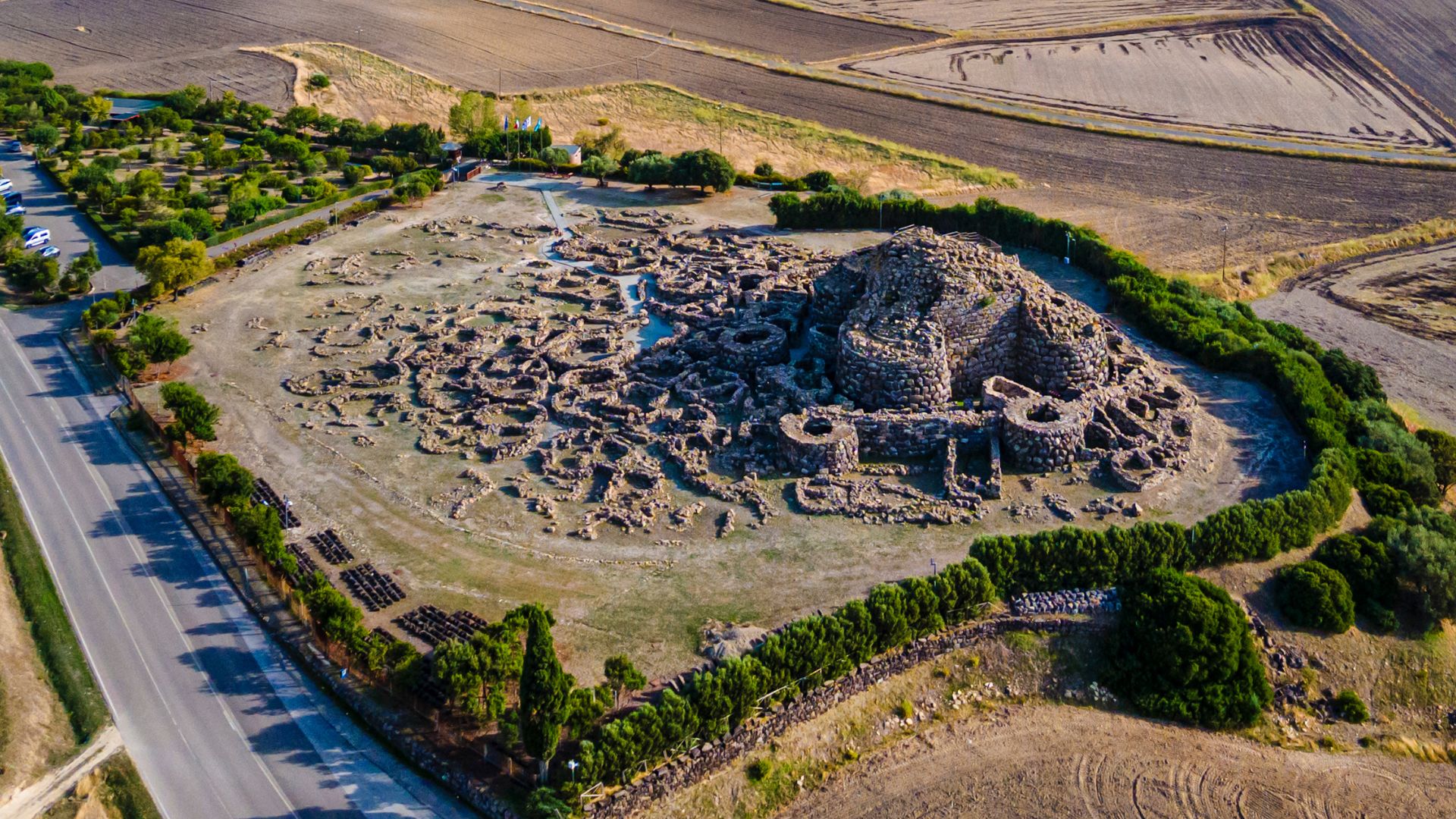 Aerial view of Su Nuraxi, a large, complex nuragic archaeological site in Sardinia, Italy, featuring a central tower and surrounding structures, enclosed by a perimeter wall and some greenery, with agricultural fields in the background and a road visible on one side.