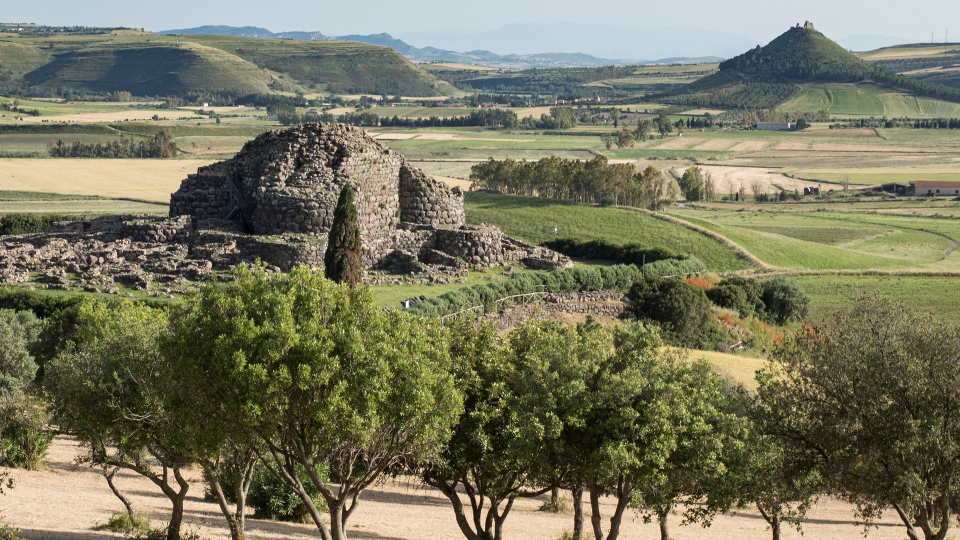 Aerial view of Su Nuraxi, a large, complex nuragic archaeological site in Sardinia, Italy, featuring a central tower and surrounding structures, enclosed by a perimeter wall and some greenery, with agricultural fields in the background and a road visible on one side. 