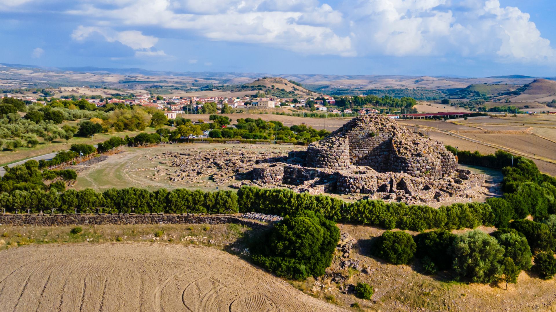 Aerial view of Su Nuraxi, a large, complex nuragic archaeological site in Sardinia, Italy, featuring a central tower and surrounding structures, enclosed by a perimeter wall and some greenery, with agricultural fields in the background and a road visible on one side. 