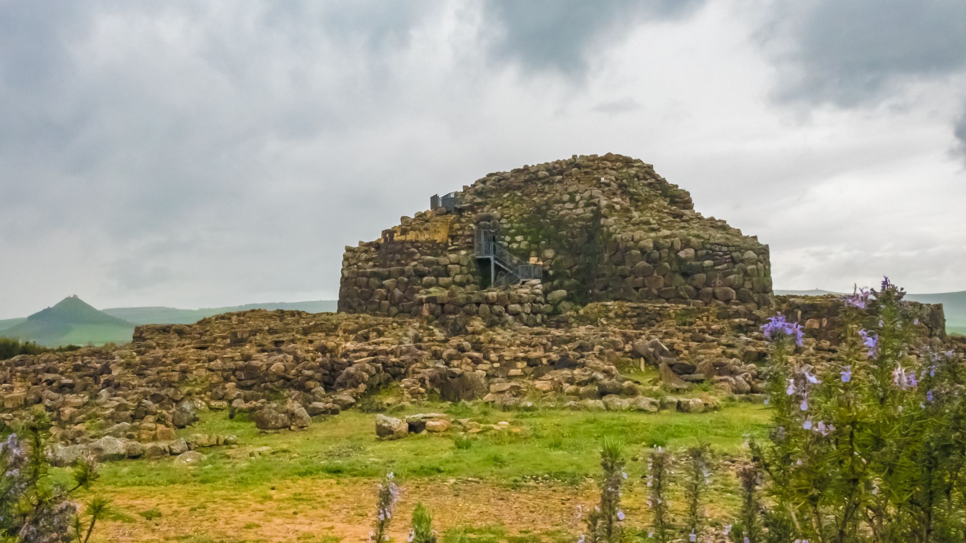 Aerial view of Su Nuraxi, a large, complex nuragic archaeological site in Sardinia, Italy, featuring a central tower and surrounding structures, enclosed by a perimeter wall and some greenery, with agricultural fields in the background.