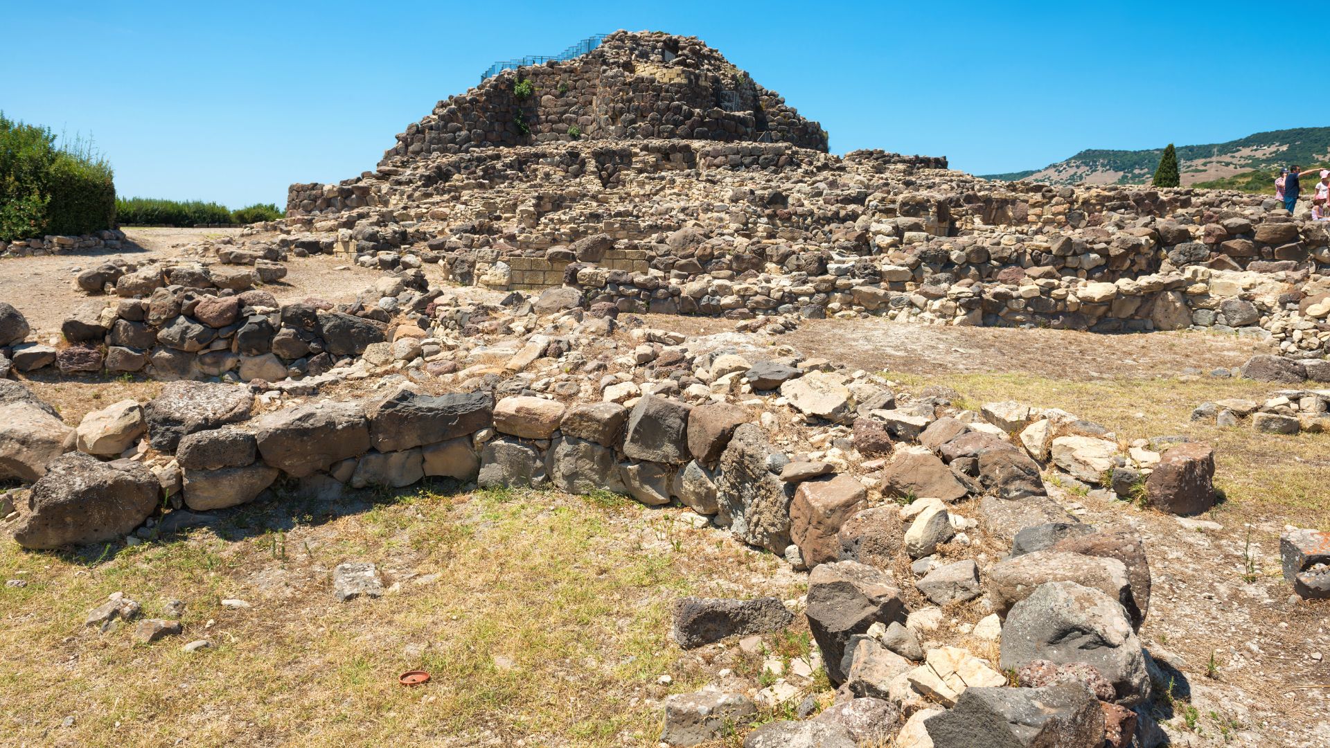 Su Nuraxi, a large, complex nuragic archaeological site in Sardinia, Italy, featuring a central tower and surrounding structures, enclosed by a perimeter wall and some greenery.
