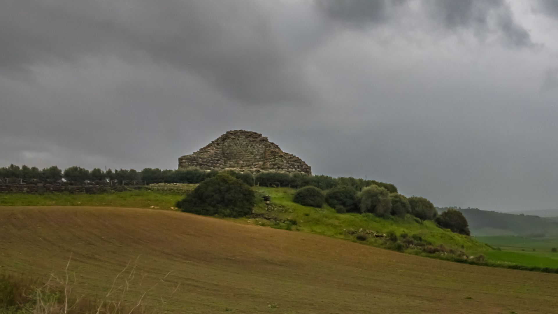 Aerial view of Su Nuraxi, a large, complex nuragic archaeological site in Sardinia, Italy, featuring a central tower and surrounding structures, enclosed by a perimeter wall and some greenery, with agricultural fields in the background.