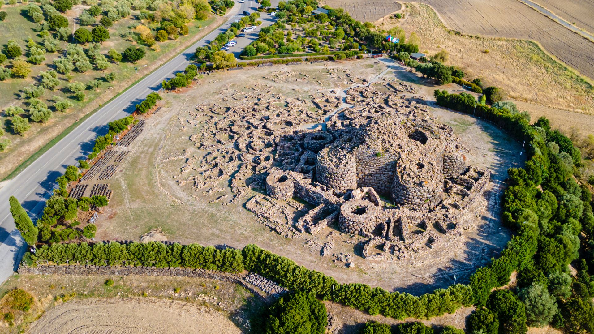 Aerial view of Su Nuraxi, a large, complex nuragic archaeological site in Sardinia, Italy, featuring a central tower and surrounding structures, enclosed by a perimeter wall and some greenery, with agricultural fields in the background and a road visible on one side. 
