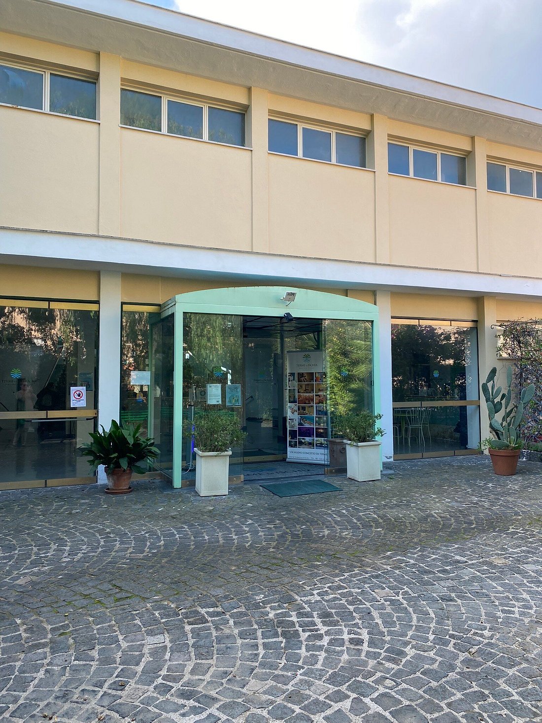 Entrance of Terme di Ischia, a spa facility in Ischia, Italy, with glass doors, potted plants, and a cobblestone walkway.