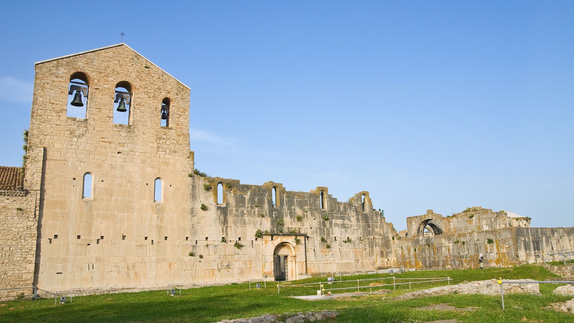 The image shows the incomplete ruins of the Abbey of Santissima Trinità in Venosa, Italy, also known as L'Incompiuta. The structure features a large, partially intact stone facade with arched openings for bells, connected to crumbling walls and archways, suggesting a rich history and architectural significance.