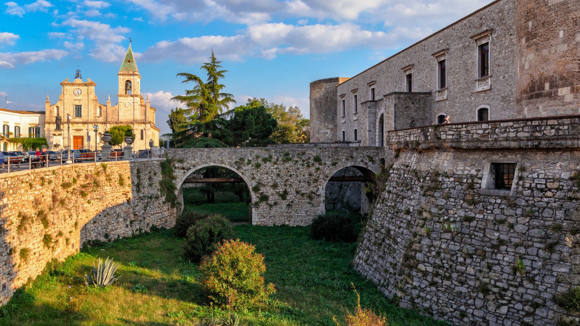 The image shows the Aragonese Castle of Venosa, Italy, featuring a stone fortress wall with arched passageways and a grassy moat. In the background, a church with a bell tower is visible, set against a blue sky with scattered clouds.