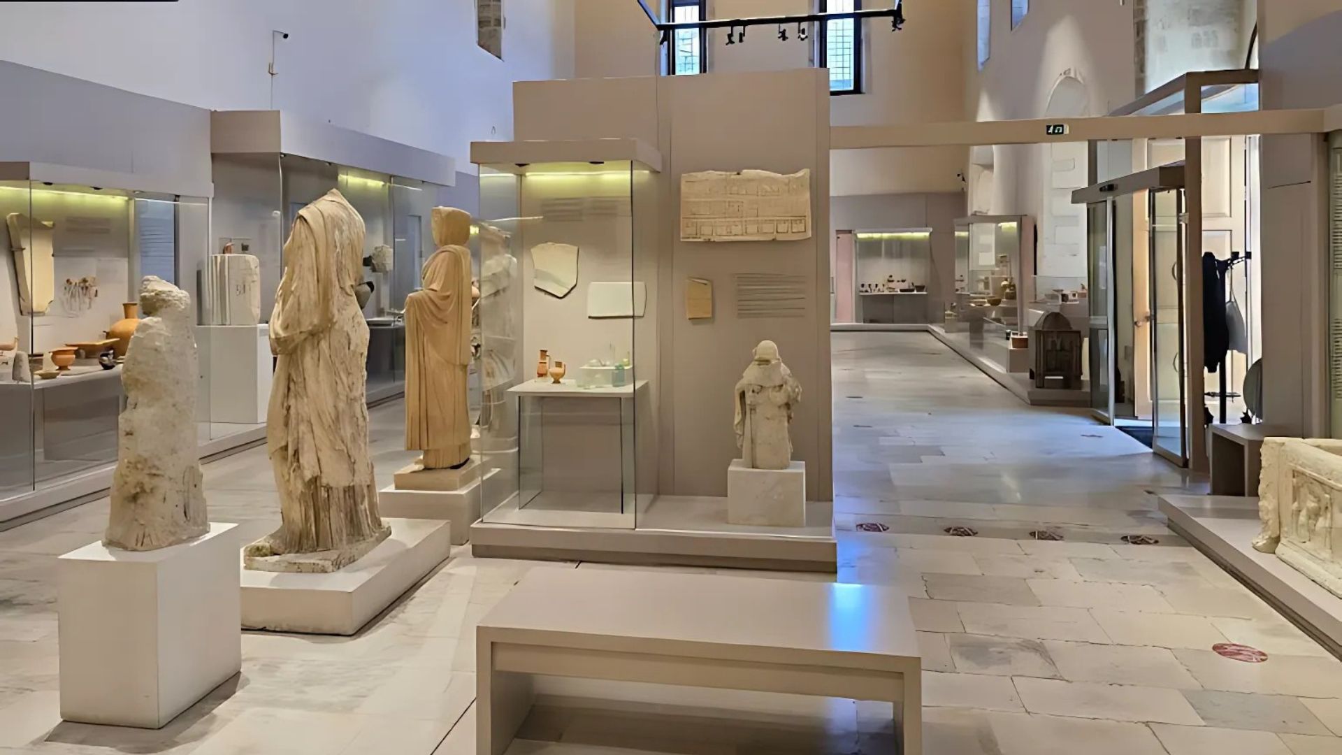 The image shows the interior of the Archaeological Museum of Rethymno, featuring a variety of ancient artifacts displayed in glass cases and on pedestals.