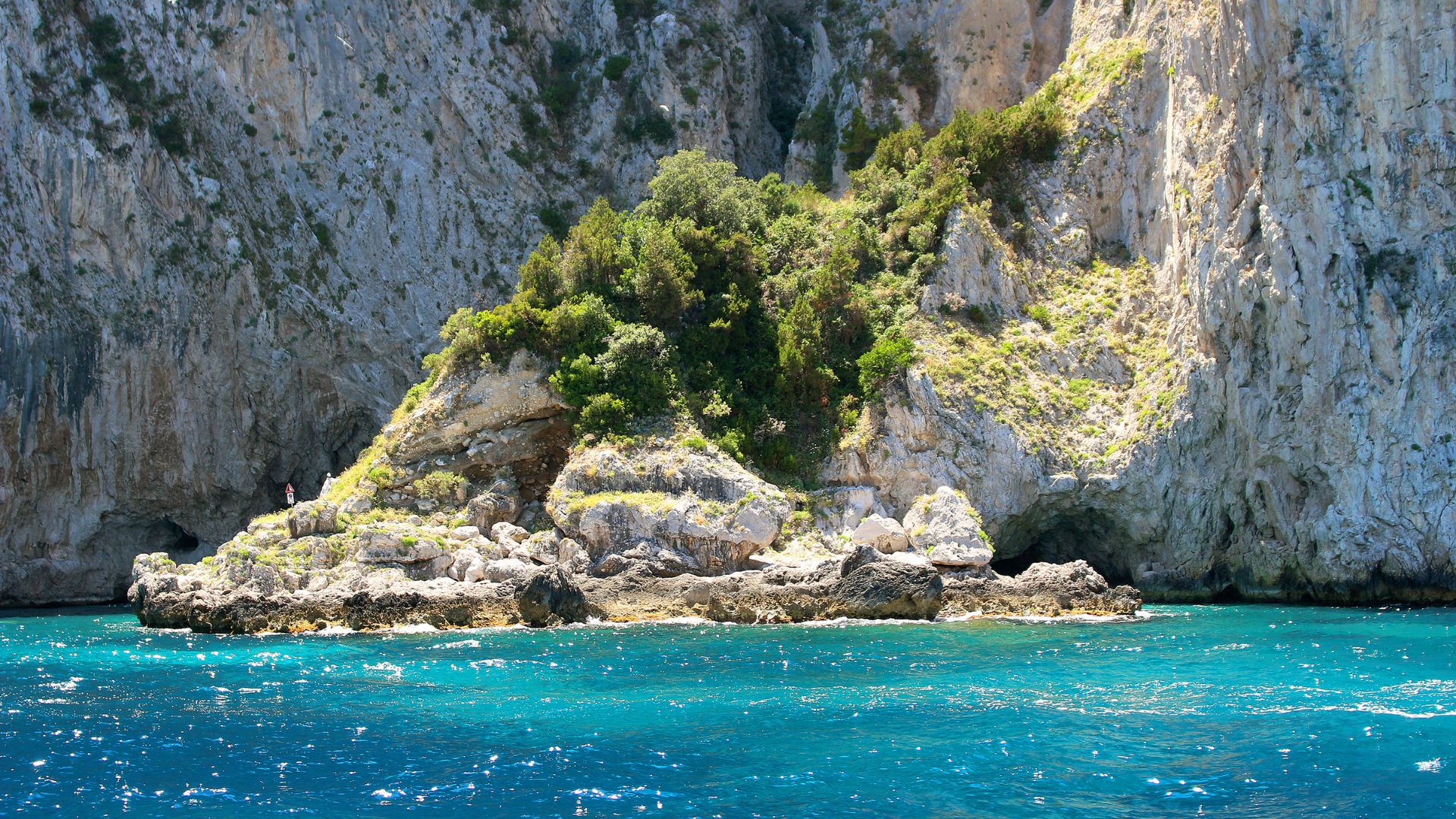 Image shows torquiese water and rocky cliff and cave of The Blue Grotto, Capri, Italy.