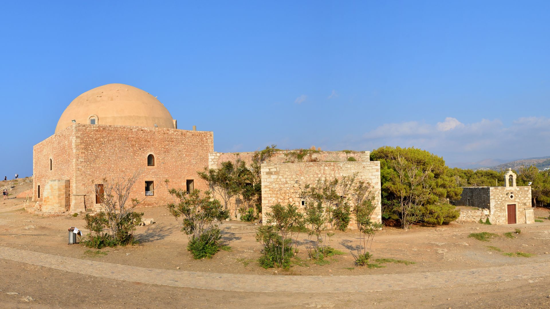 The Ibrahim Han Mosque, a stone building with a large dome and small windows, stands inside the Fortezza of Rethymno in Crete, surrounded by sparse vegetation and a dirt path, under a clear blue sky.