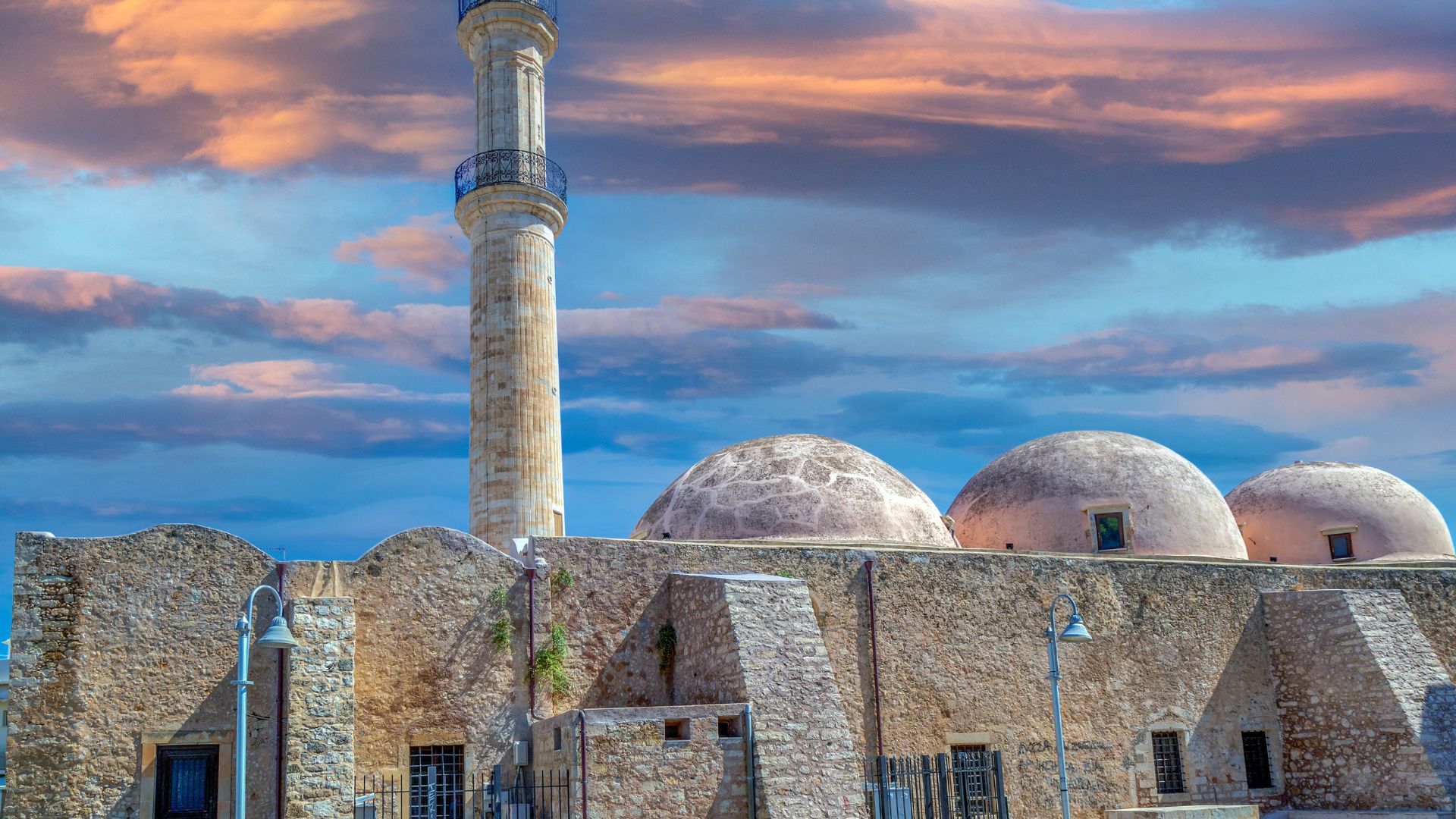The Neratze Mosque in Rethymno, Crete, featuring a tall minaret and domed roofs, set against a cloudy sky.