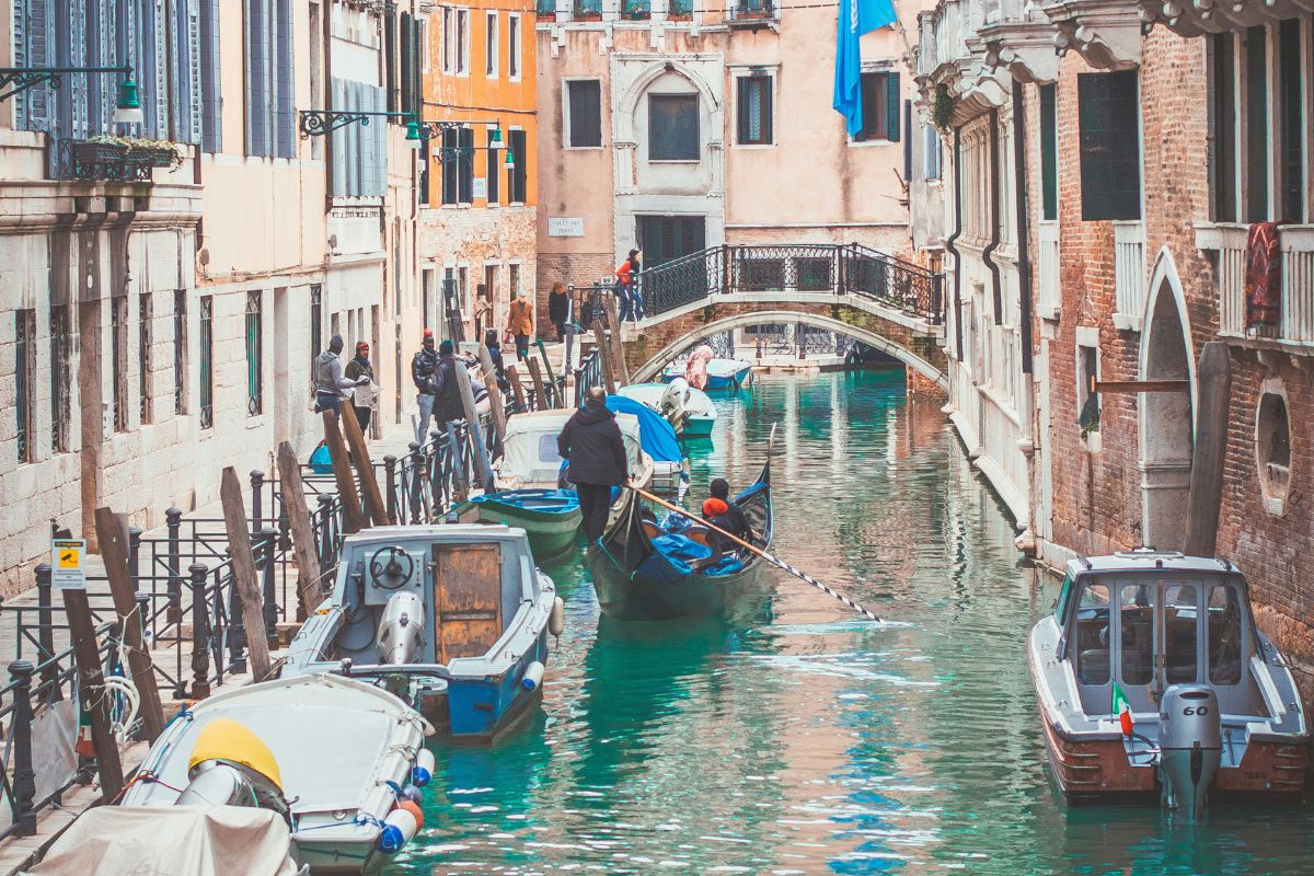 Boats and gondolas navigate a canal in Venice, surrounded by colorful buildings and a bridge. The image captures the characteristic Venetian scene with boats on the water and the city's architecture in the background.