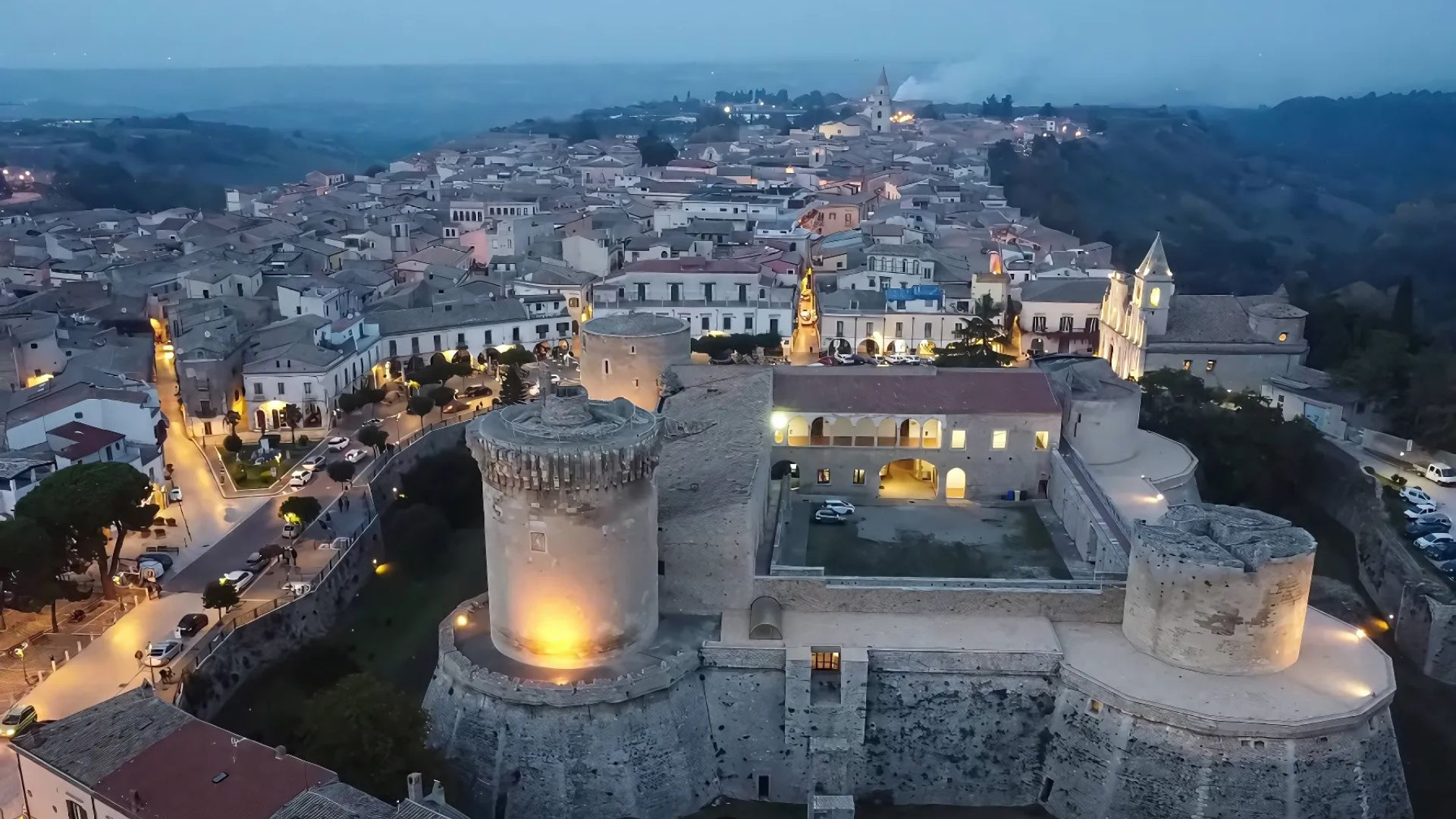 Aerial view of the illuminated Aragonese Castle (Castello del Balzo) in Venosa, Italy, with the town's buildings and landscape visible in the background.