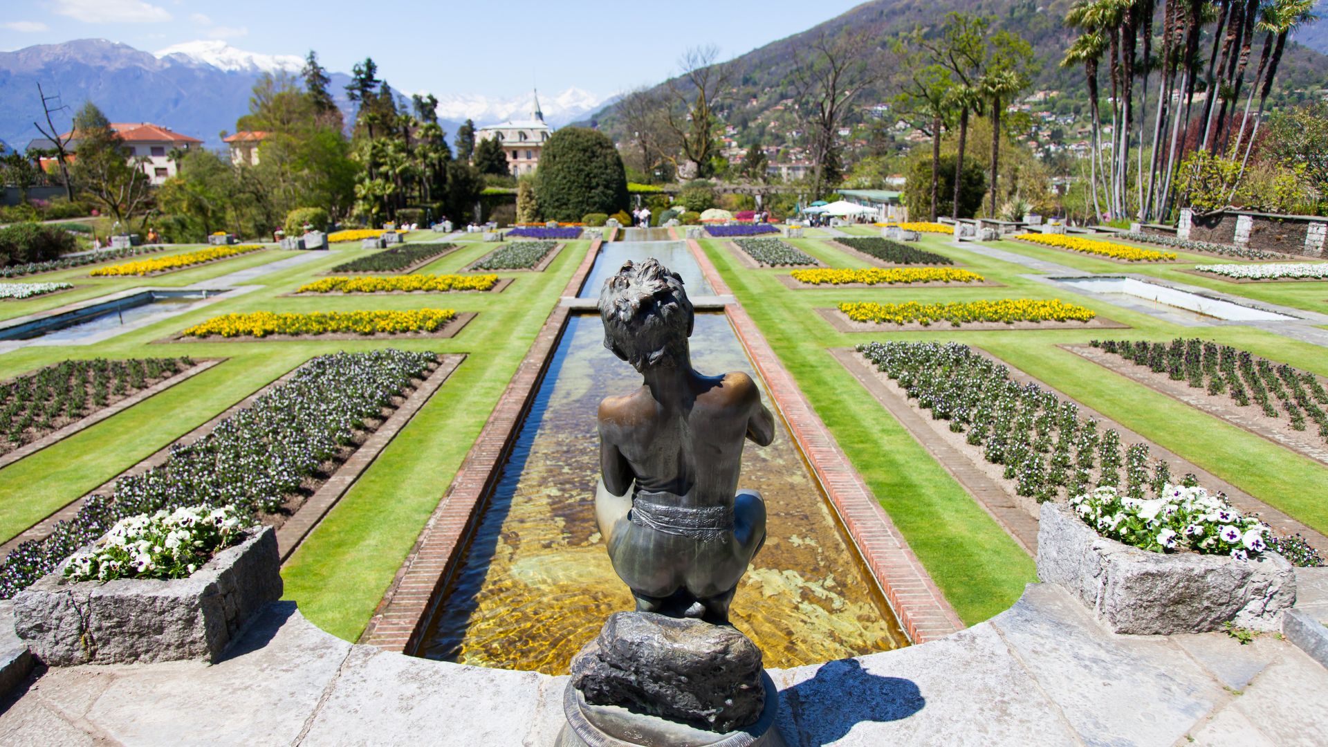 A statue overlooks the symmetrical and colorful gardens of Villa Taranto, with mountains visible in the distance.