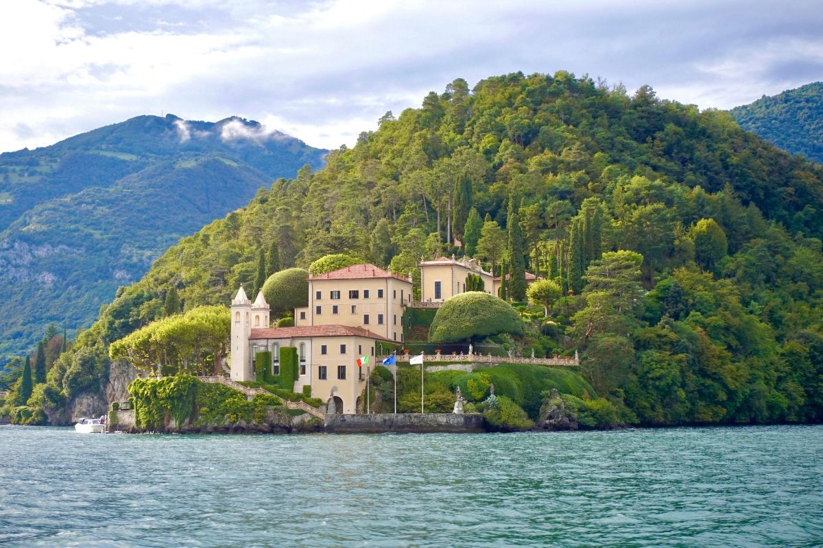 Villa del Balbianello on Lake Como, a beige building with a tower surrounded by lush greenery and water, with mountains in the background.