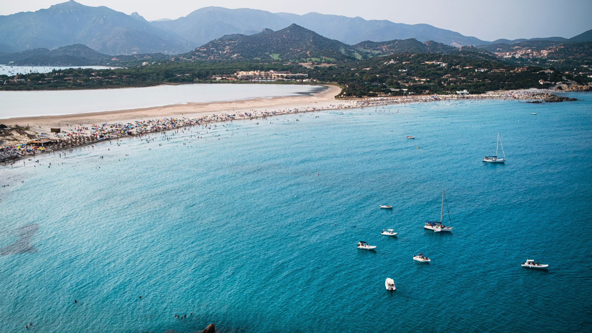 Aerial view of blue water, white sand beaches, green mountains, boats, and beach goers at Villasimius, Sardinia, Italy.