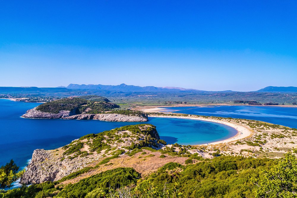 Clear blue waves gently lapping against the soft sand of Voidokilia Beach under a bright summer sky.