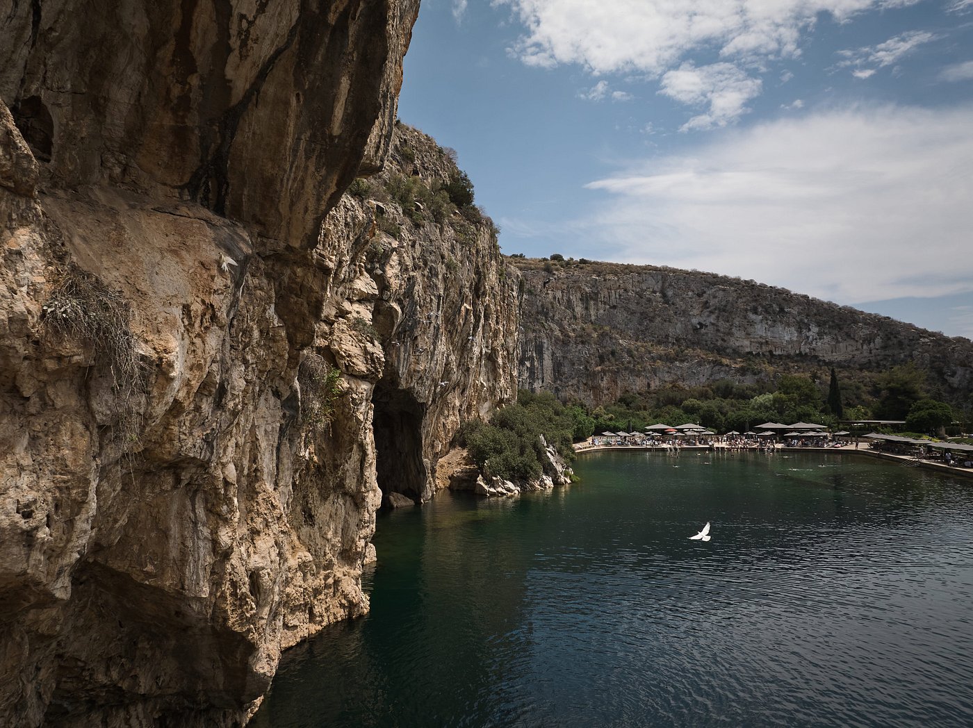 Rocky cliffs and calm waters at Vouliagmeni Lake with a view of a cave and nearby visitors area in Greece.