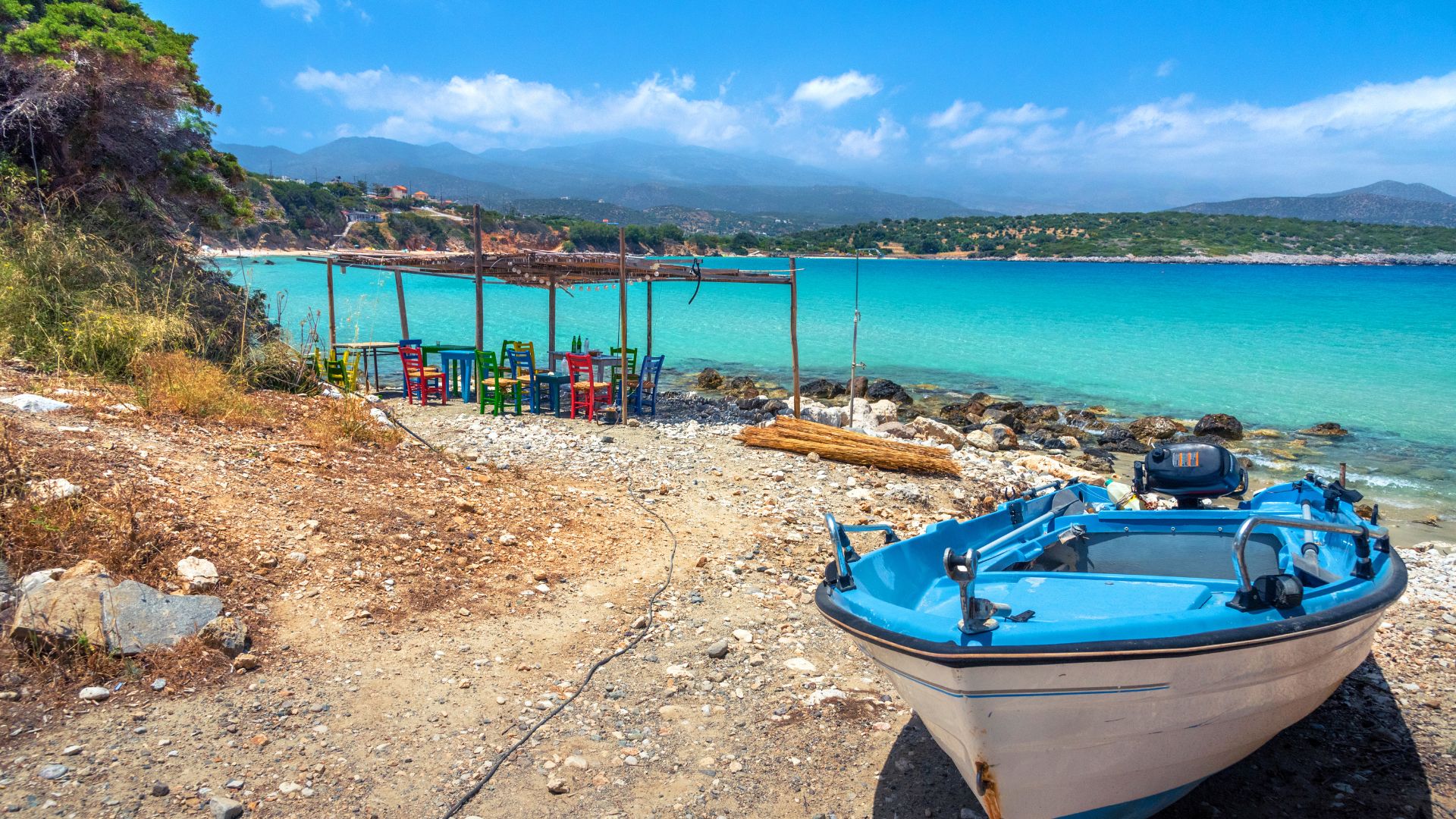 A scenic view of Voulisma Beach in Crete, featuring turquoise waters, a sandy shore with a boat, and beachside seating under shaded structures.