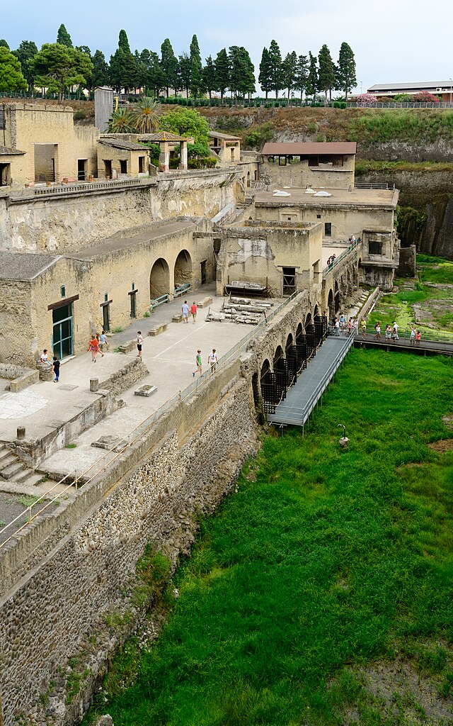 Ruins of the Herculaneum boat houses with arched structures and visitors walking on elevated paths.